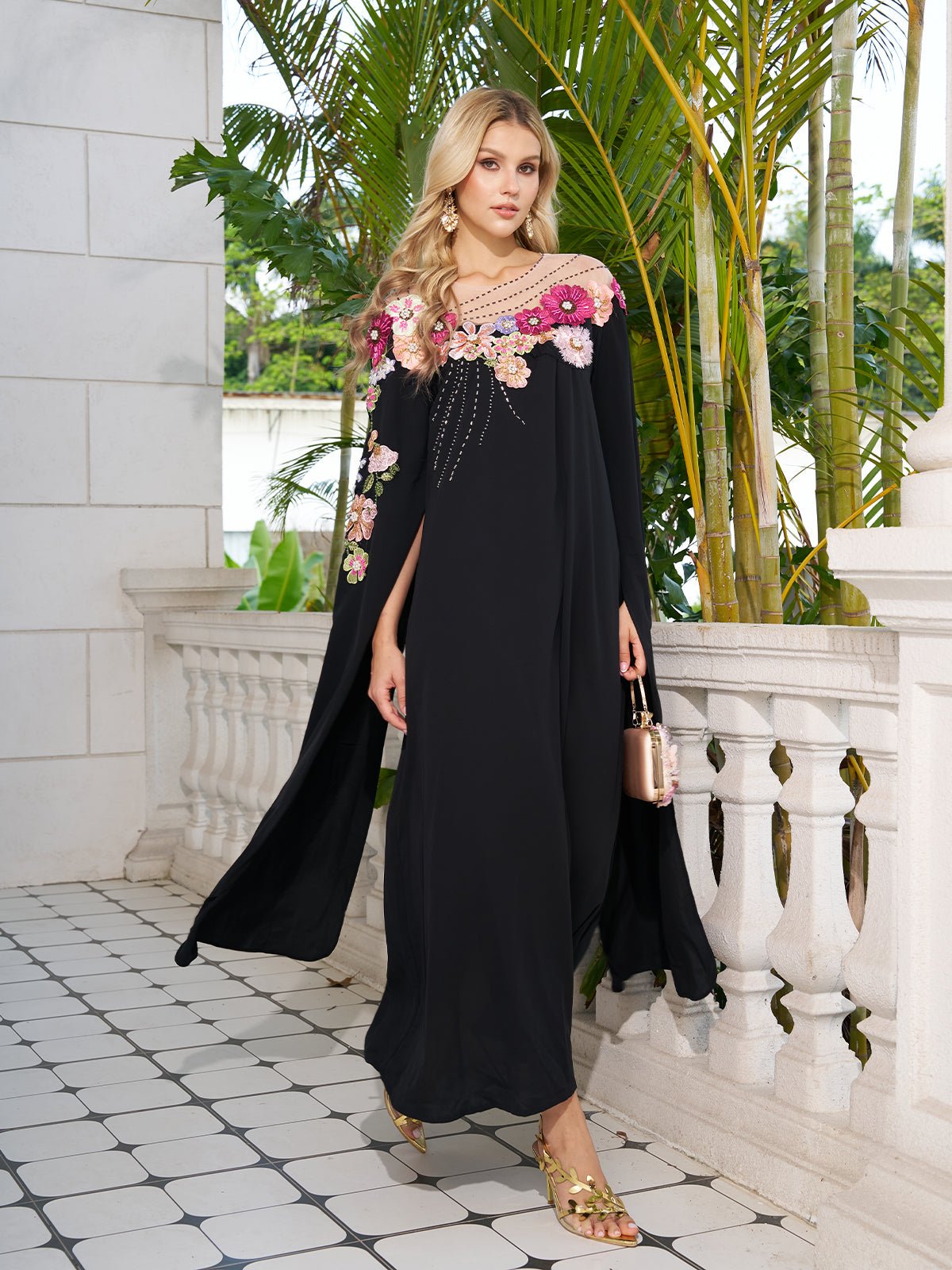A woman wears the Social Queen Hand-Embroidered Floral Cape Gown—black with pink floral accents—while standing on a tiled balcony with white railings and tropical plants behind her.