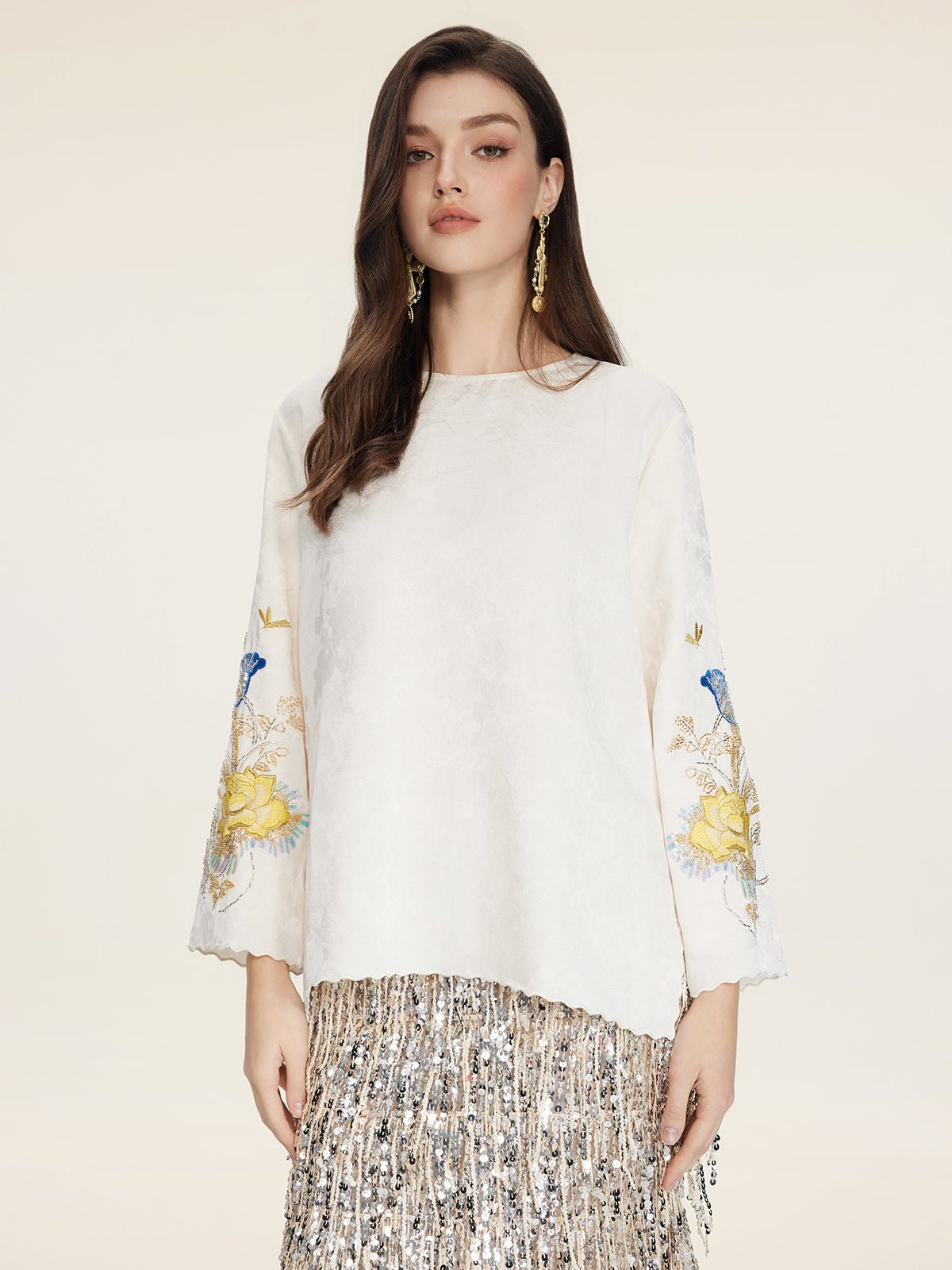 A woman models the Social Queen Hand-Embroidered Floral Jacquard Pullover with floral sleeve motifs, paired with a silver fringed skirt, against a plain background.