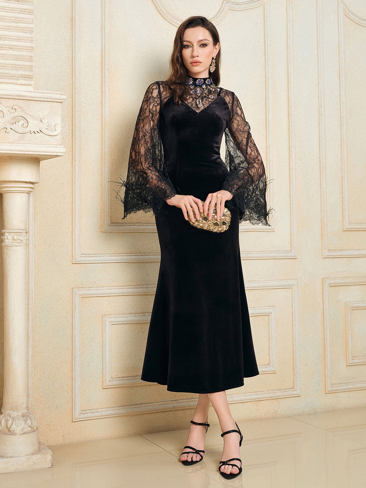 A woman wearing the Social Queen Hand-Sewn Crystal Velvet Feather Mermaid Gown holds a gold clutch, standing indoors against a decorative cream-colored wall.