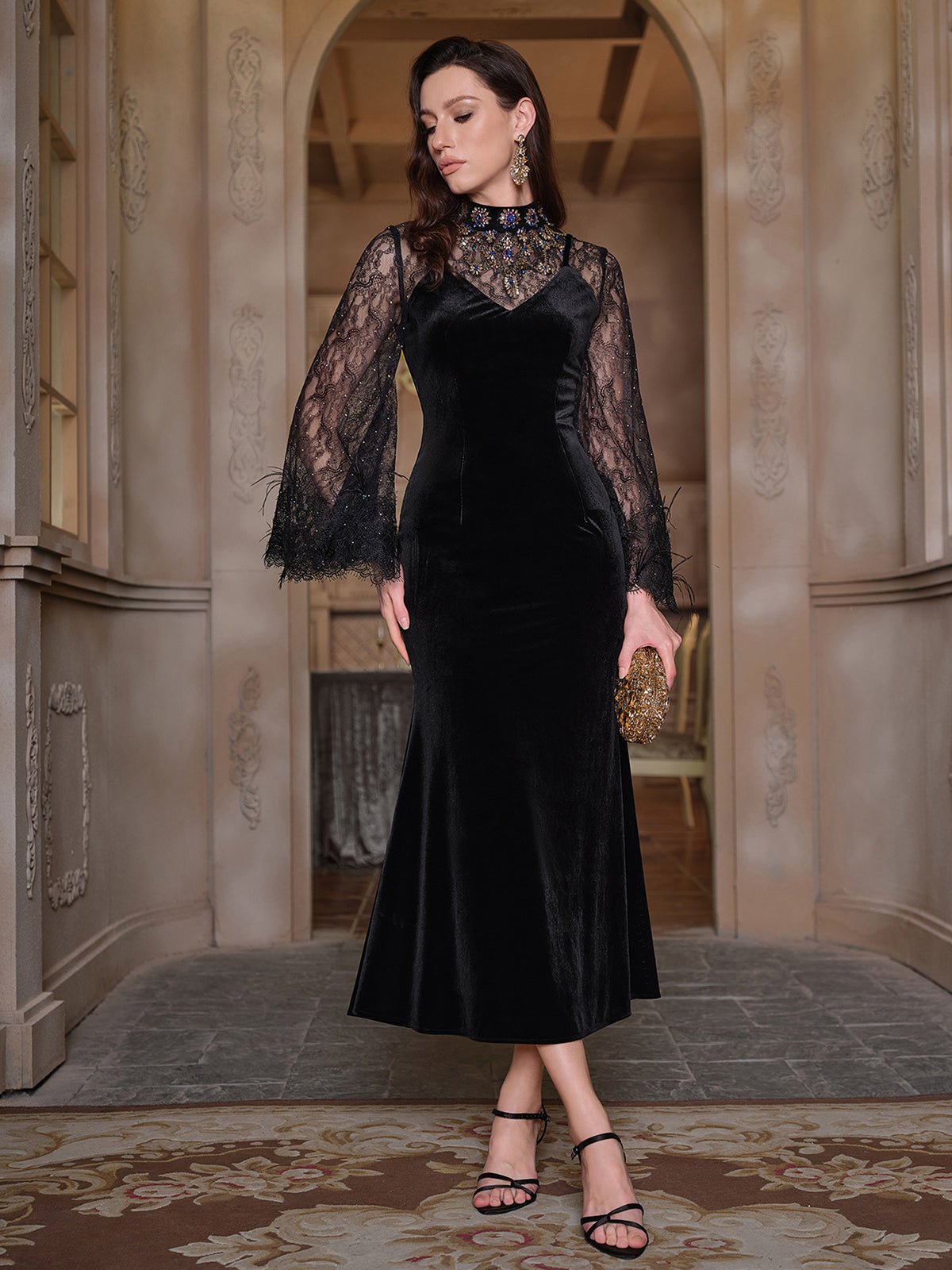 A woman stands indoors wearing the Social Queen Hand-Sewn Crystal Velvet Feather Mermaid Gown with sheer lace-detailed sleeves, an embellished neckline, black sandals, and a small clutch.
