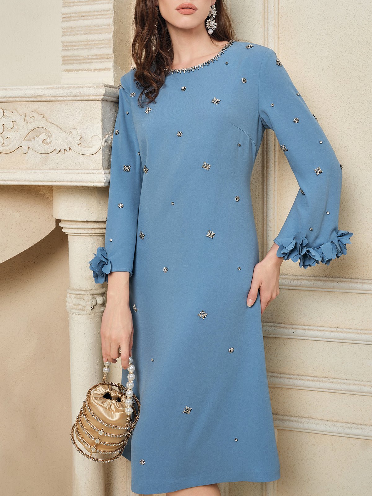A woman wearing the Social Queen Handcrafted Beaded Floral A-Line Dress stands beside a mantel, holding a gold handbag with a beaded handle.