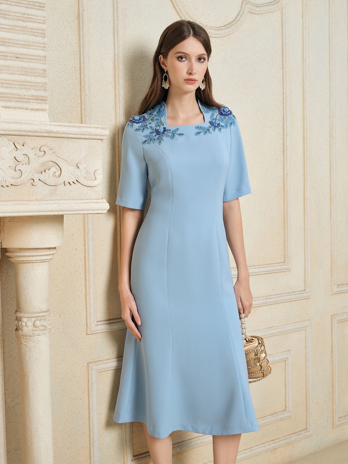 A woman stands indoors wearing the Social Queen Handcrafted Beaded Floral Dress with hand-embroidered 3D floral appliqués on the shoulders, while holding a small beige handbag.