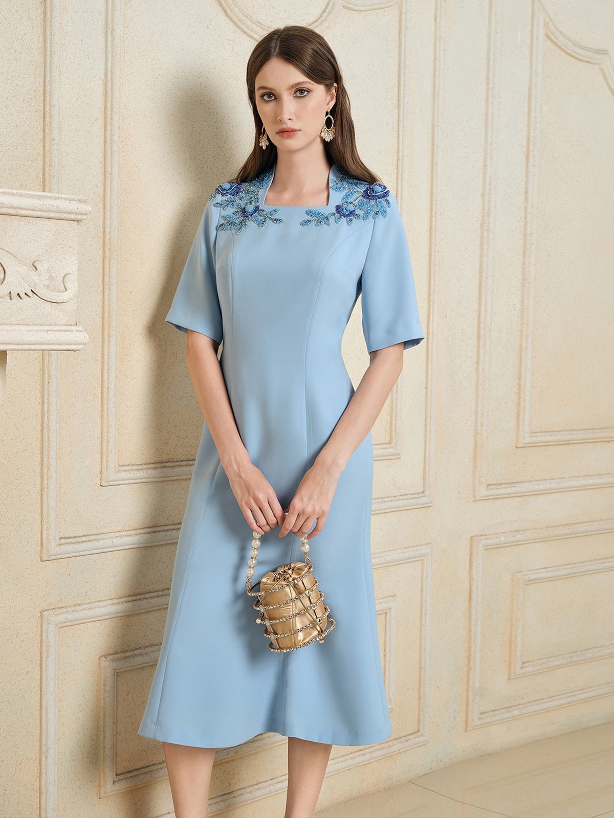 A woman wearing the Social Queen Handcrafted Beaded Floral Dress stands against a beige wall, holding a small beaded handbag.