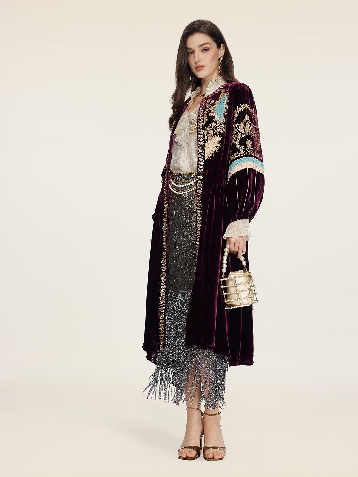 A woman radiates vintage glamour in the Social Queen Handcrafted Embroidered Velvet Jacket over a white blouse and sparkly fringed skirt, holding a metallic handbag and wearing heels against a plain backdrop.