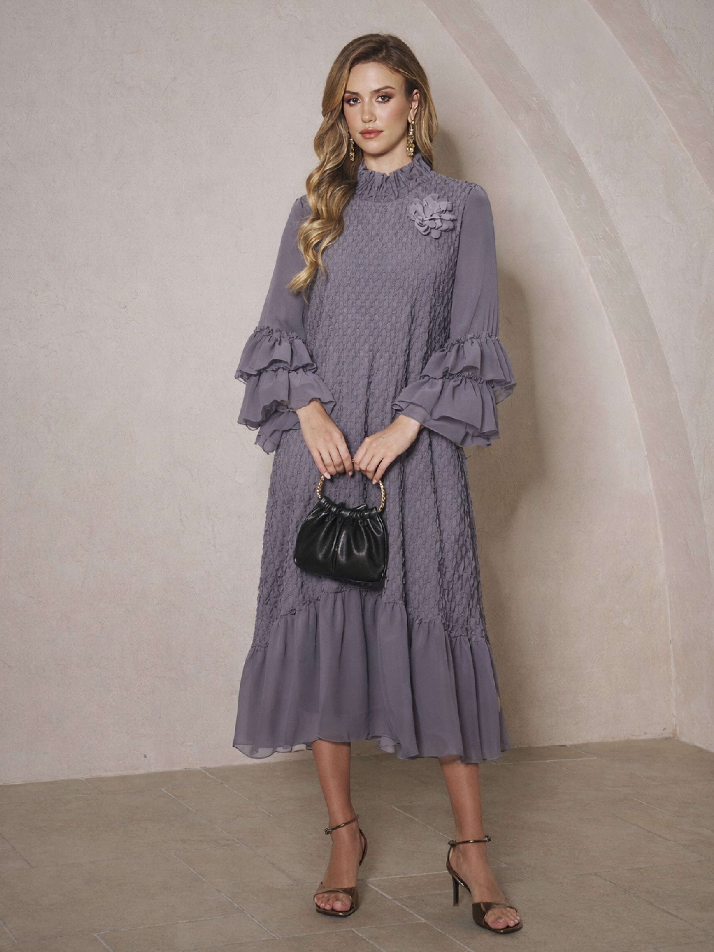 A woman holds a black purse while wearing the Social Queen Floral Appliqué Embroidered Ruffle Dress, featuring long sleeves and delicate details, standing against a beige textured wall.