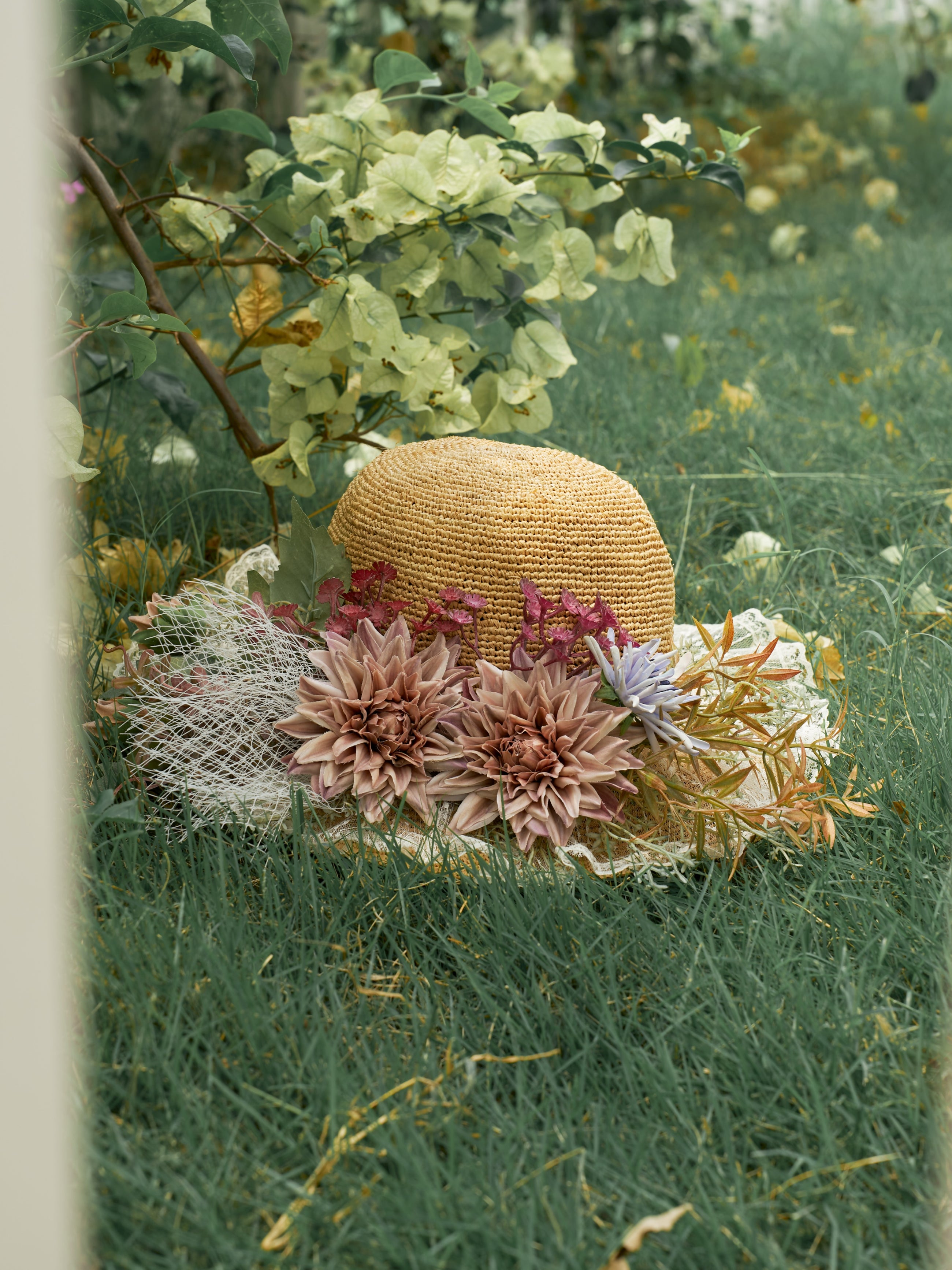 The Social Queen 3D Dahlia Raffia Wide-Brim Hat, adorned with large faux flowers and netting, rests on grass among leafy plants.