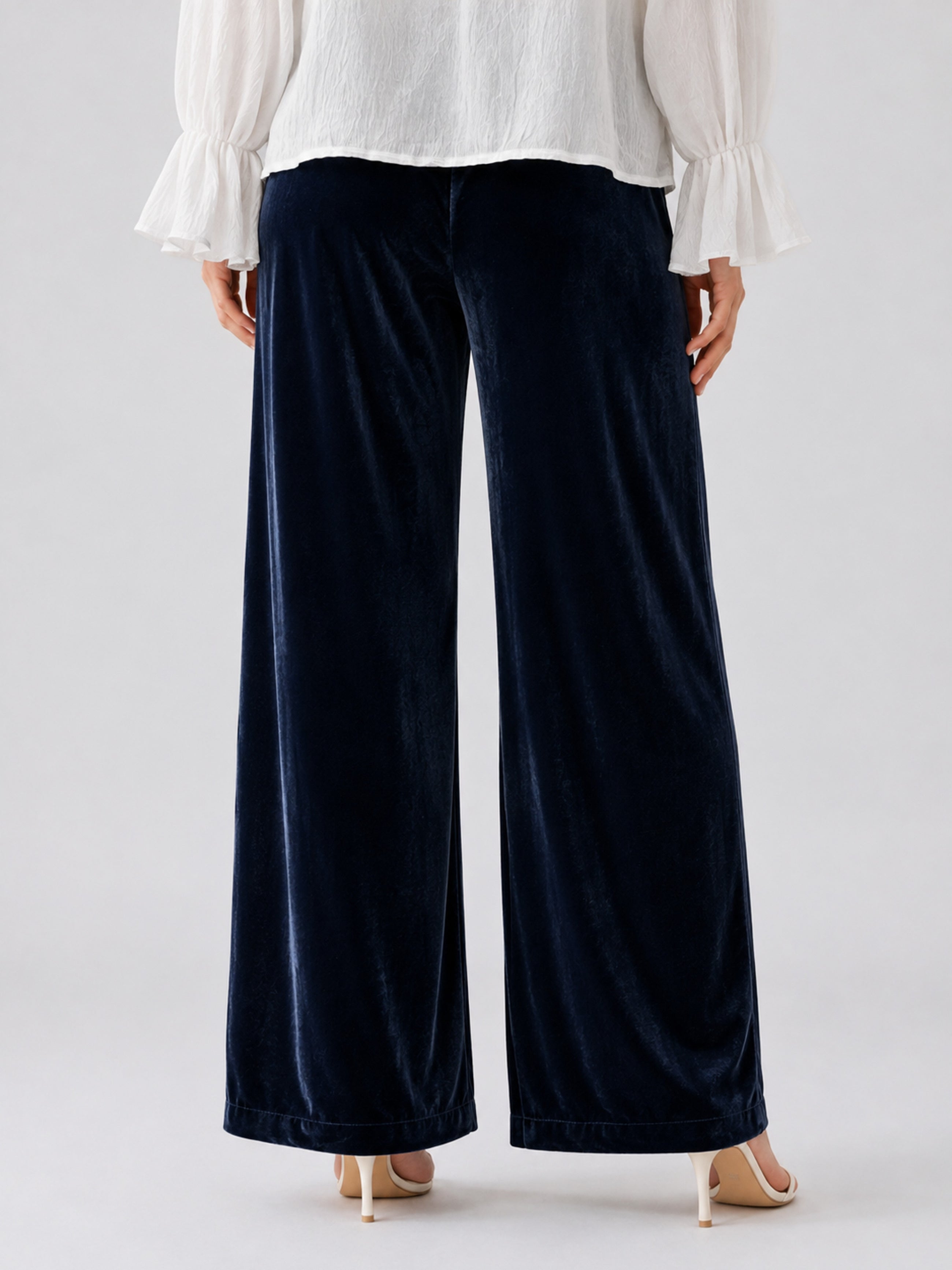 A person is shown from the back wearing Social Queen’s Silk Velvet Wide-Leg Pants in navy blue, paired with a white long-sleeve blouse featuring ruffled cuffs and beige heeled sandals.