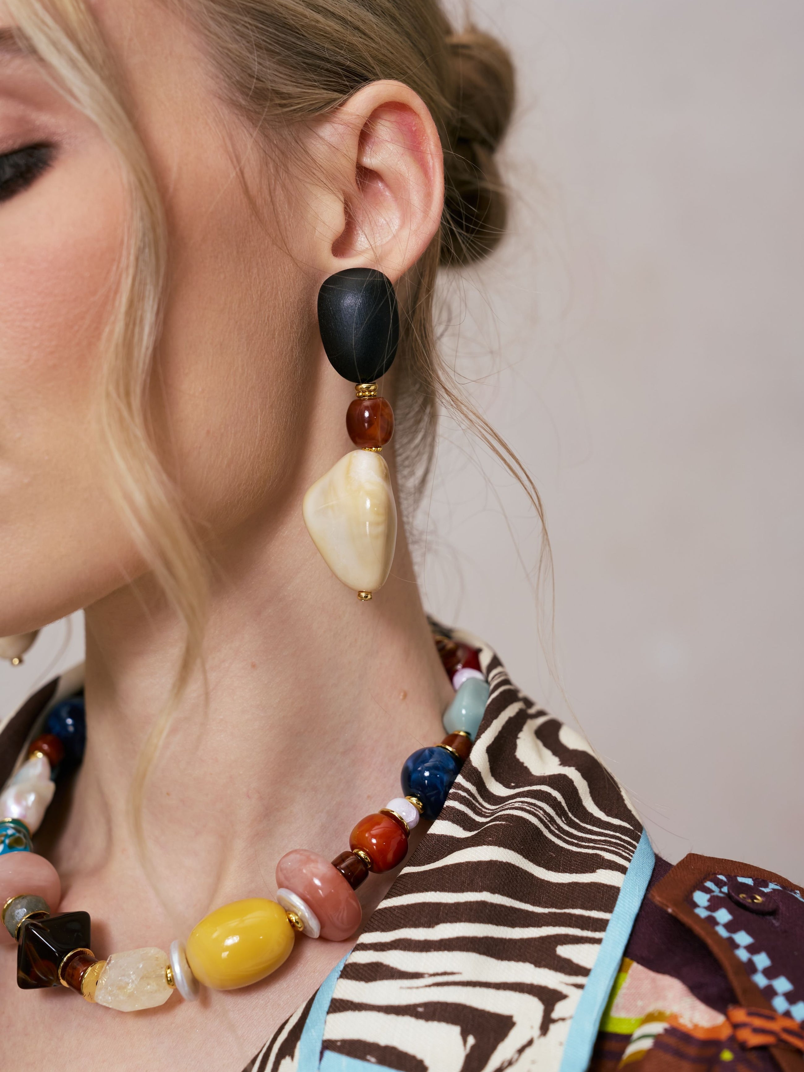 Close-up of a woman wearing the Social Queen Multicolor Beaded Drop Earrings and a beaded necklace. She is dressed in a patterned brown, white, and blue top. Only her ear, neck, and part of her face are visible, emphasizing the intricate bohemian statement earrings from Social Queen.