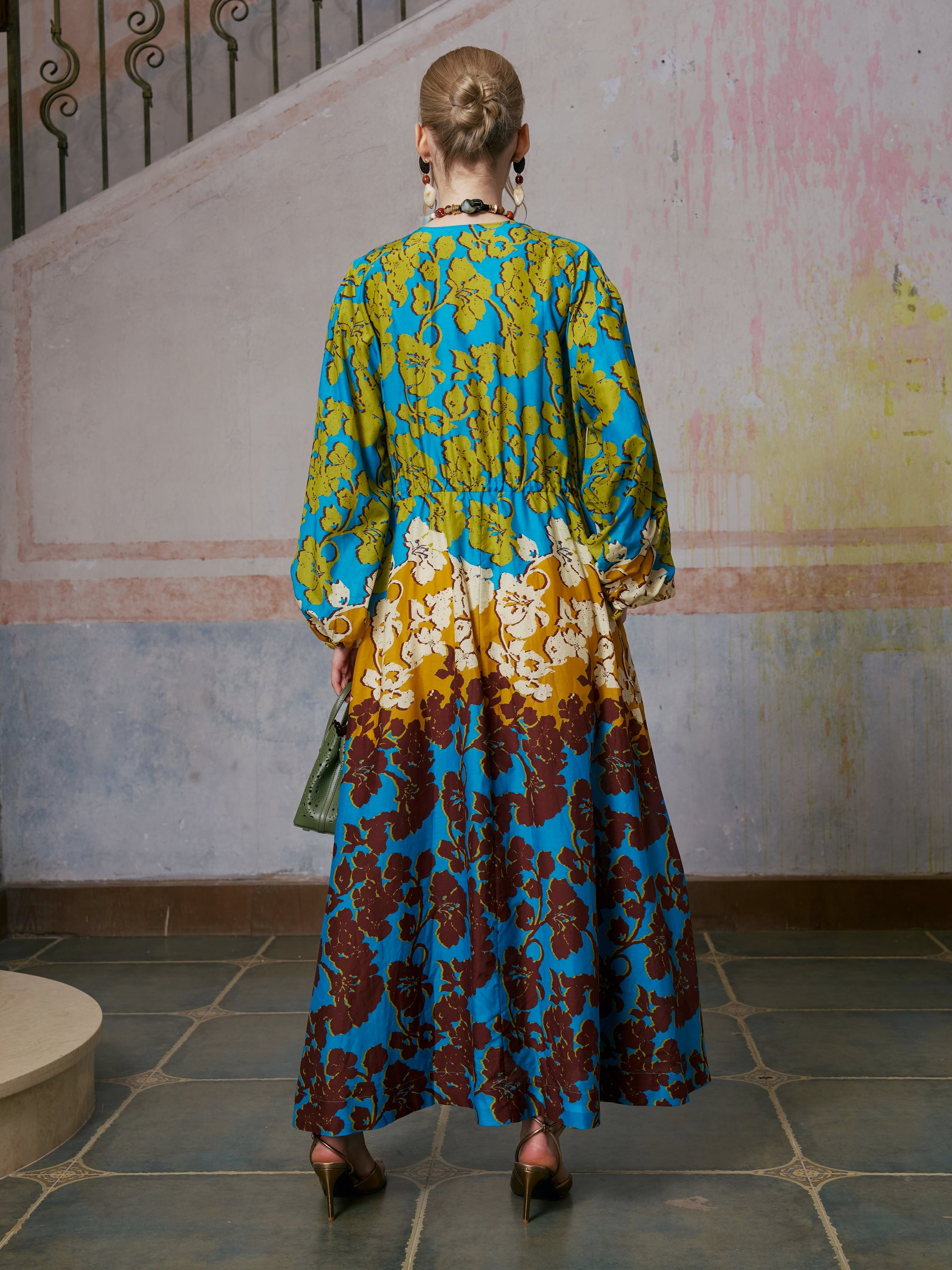 A woman stands with her back to the camera, wearing the Social Queen Contrast Floral Print Silk Cotton Dress—a long-sleeved, ankle-length piece in blue, yellow, and maroon tones. Styled with heels and statement earrings, this elegant resort-ready dress is perfect for effortless sophistication.