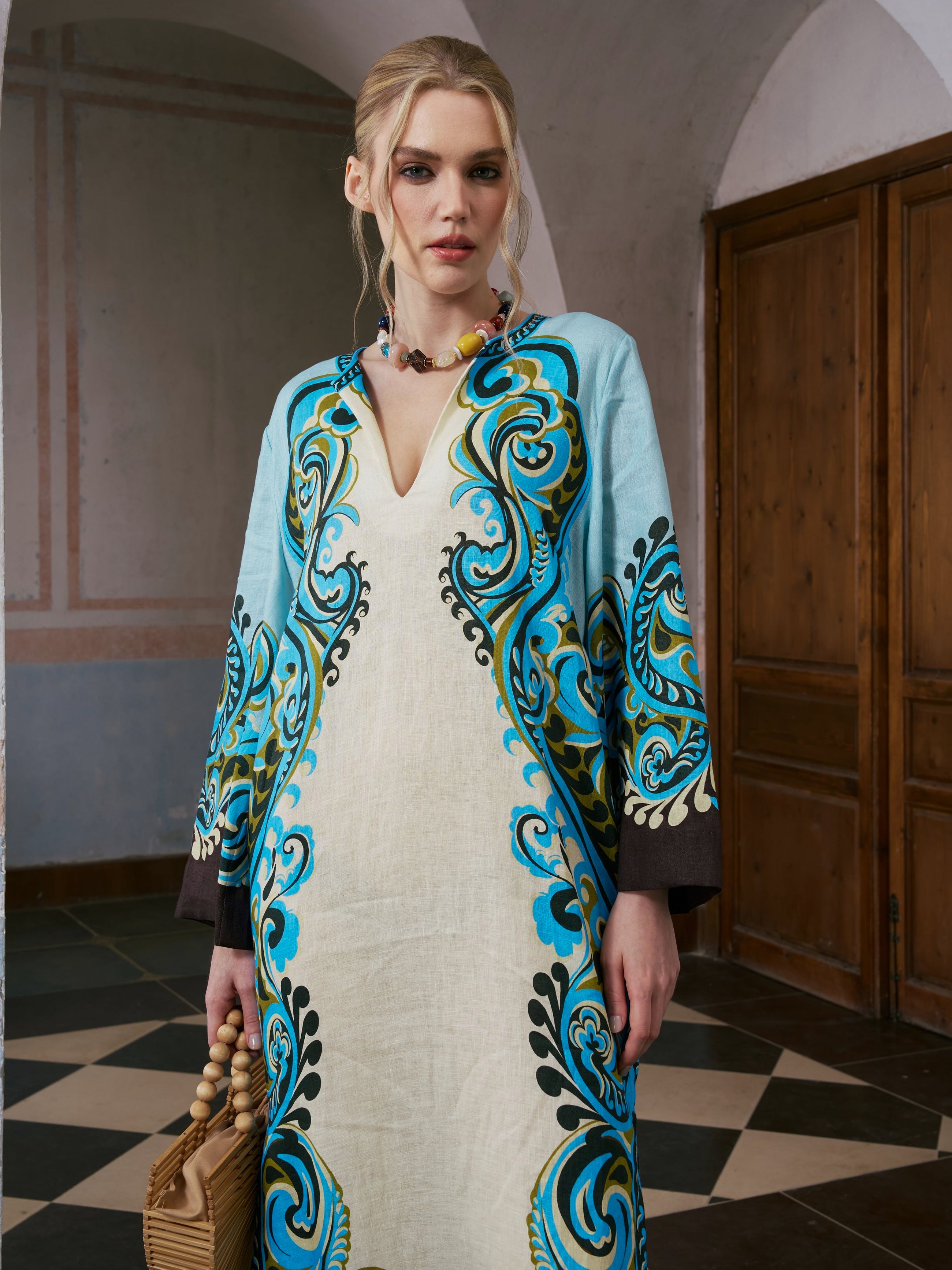 A woman with blonde hair tied back stands indoors, holding a wooden handbag and wearing the Paisley Print Linen Dress by Social Queen, featuring blue, black, and turquoise designs in a long, robe-style silhouette. She completes her look with a beaded necklace.