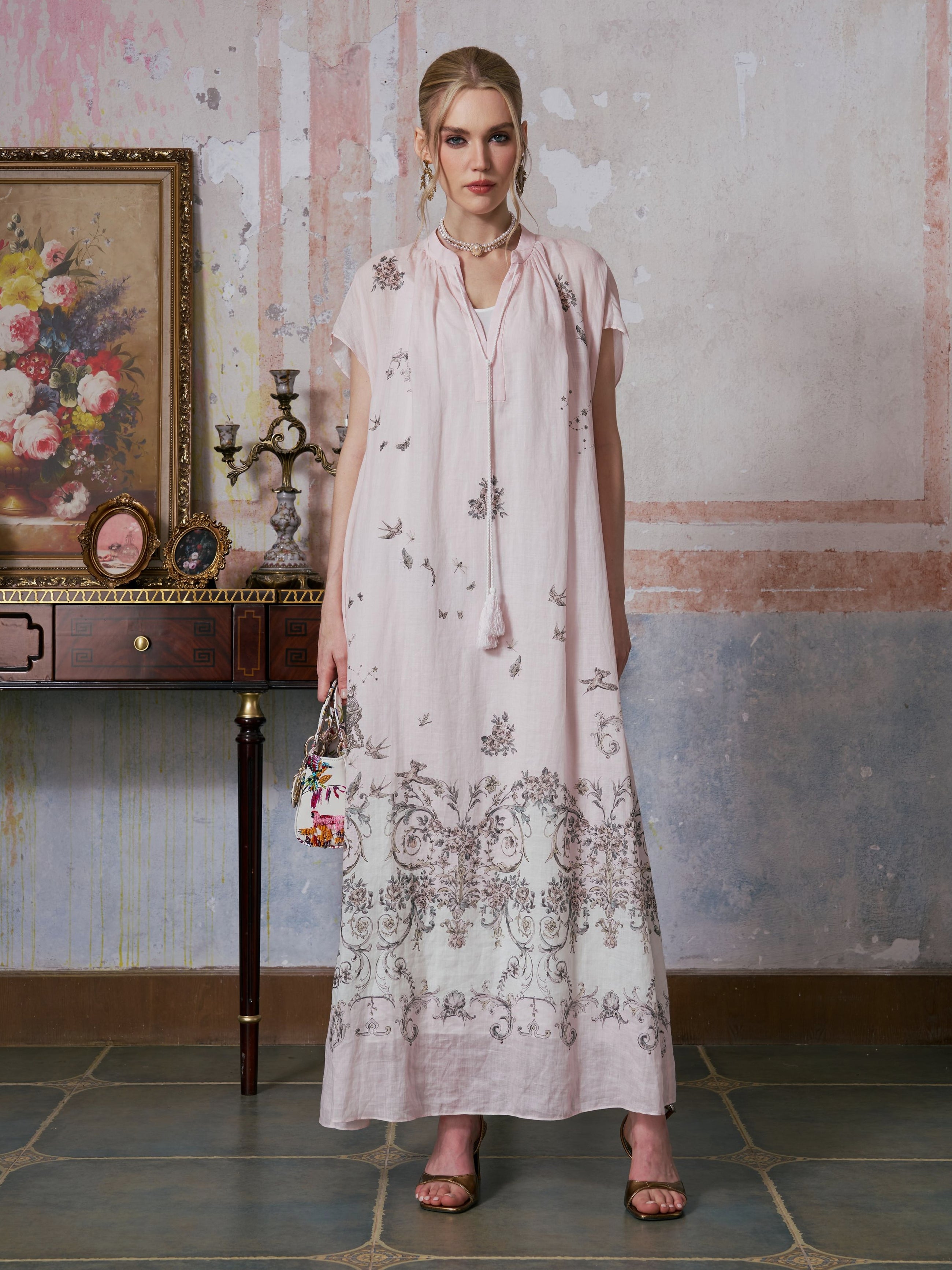 A woman stands indoors in front of a wall, holding a small handbag, wearing the Social Queen Baroque Garden Print Ramie Dress (Includes Slip Dress), with a decorative table and floral painting beside her.