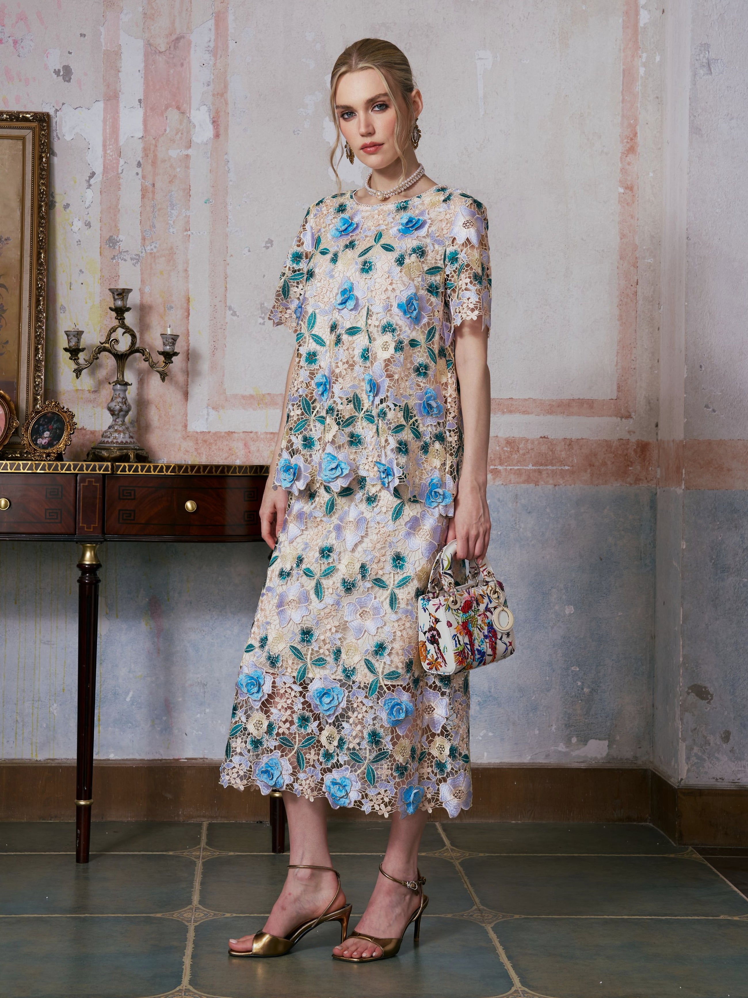 A woman stands indoors in front of a textured wall, wearing the Social Queen 3D Floral Cutwork Lace Skirt paired with a floral lace dress featuring blue flowers. She holds a small handbag and completes her look with gold sandals.