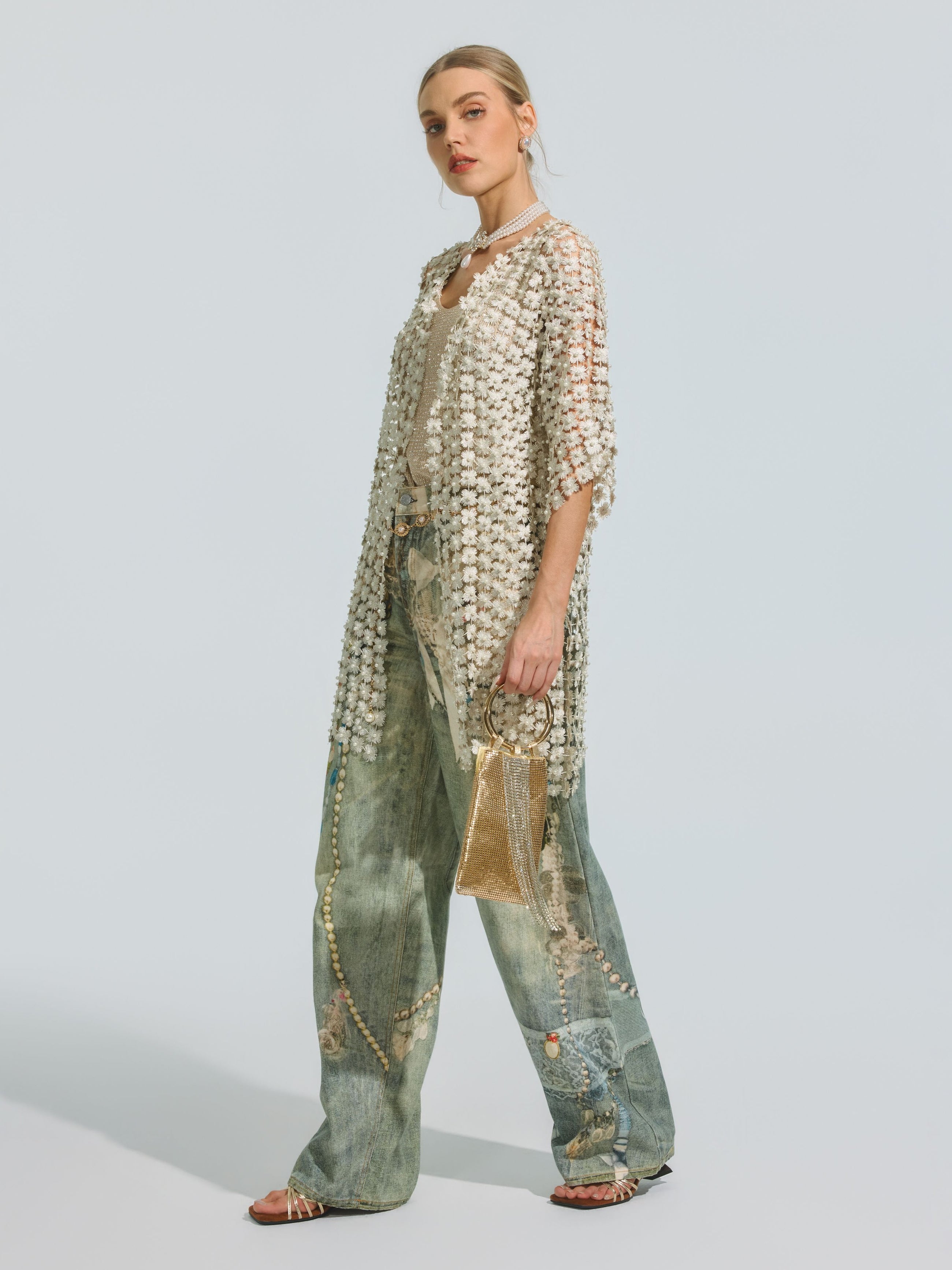 Wearing the Social Queen Pearl Daisy Cutwork Long Cardigan, adorned with delicate pearl embellishments over a tank and wide-leg jeans, and accessorized with a small gold purse, the person stands against a plain background.