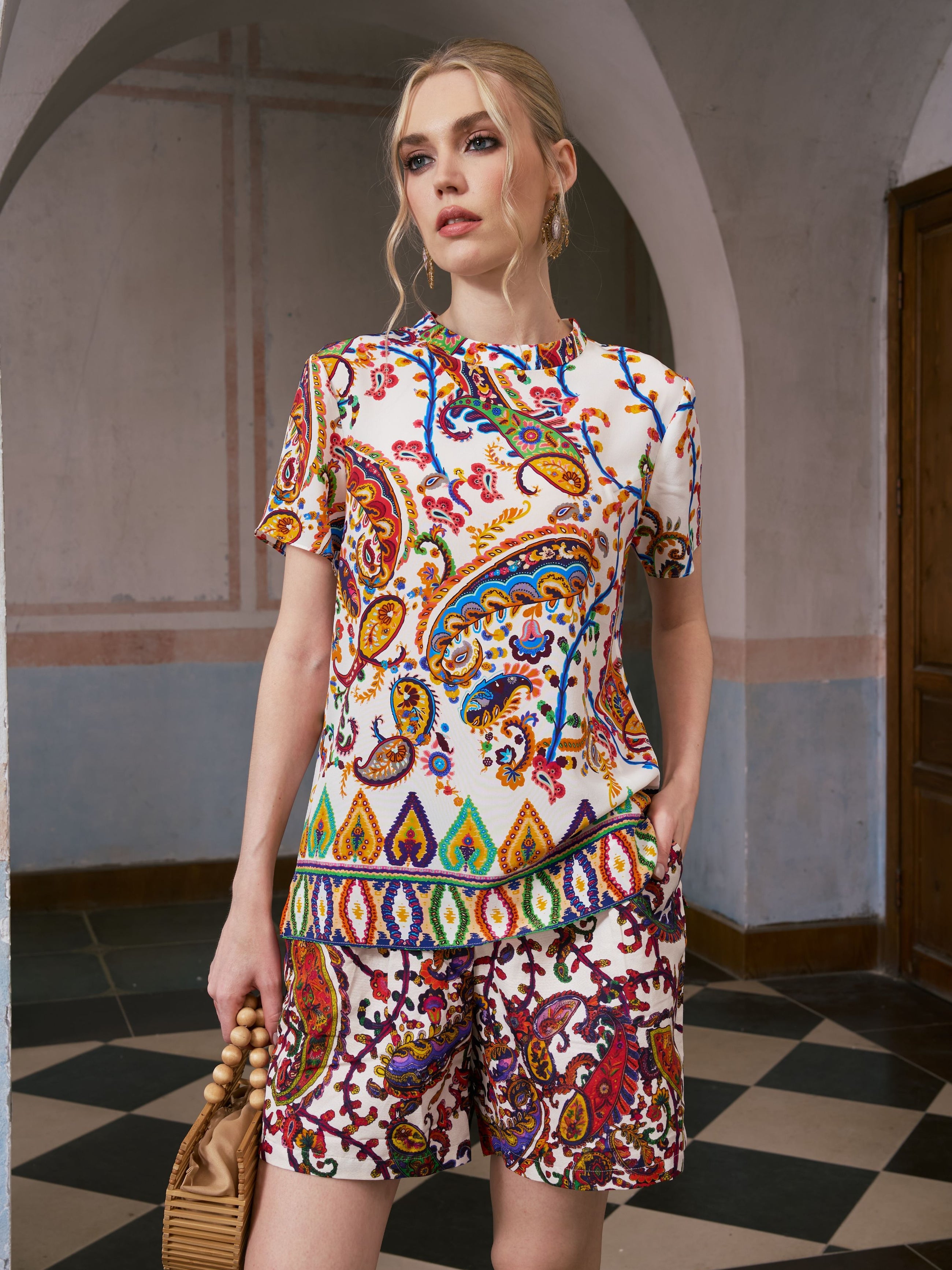 A woman stands indoors on a checkered floor, holding a wooden beaded handbag and wearing the Social Queen Paisley Garden Print Silk Top with matching shorts—a vibrant, effortless choice for resort wear.