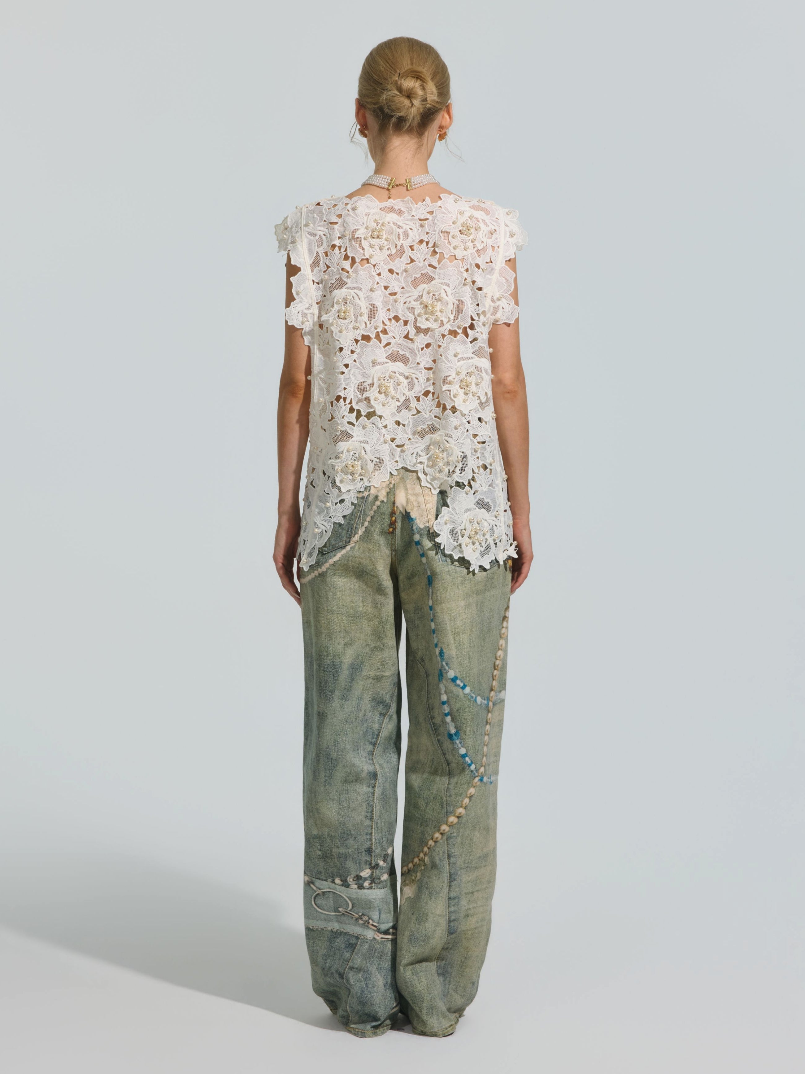 A woman stands facing away, wearing the Social Queen Pearl Rose Guipure Lace Top paired with loose-fitting, faded jeans adorned with decorative beads, chains, and subtle pearl details.