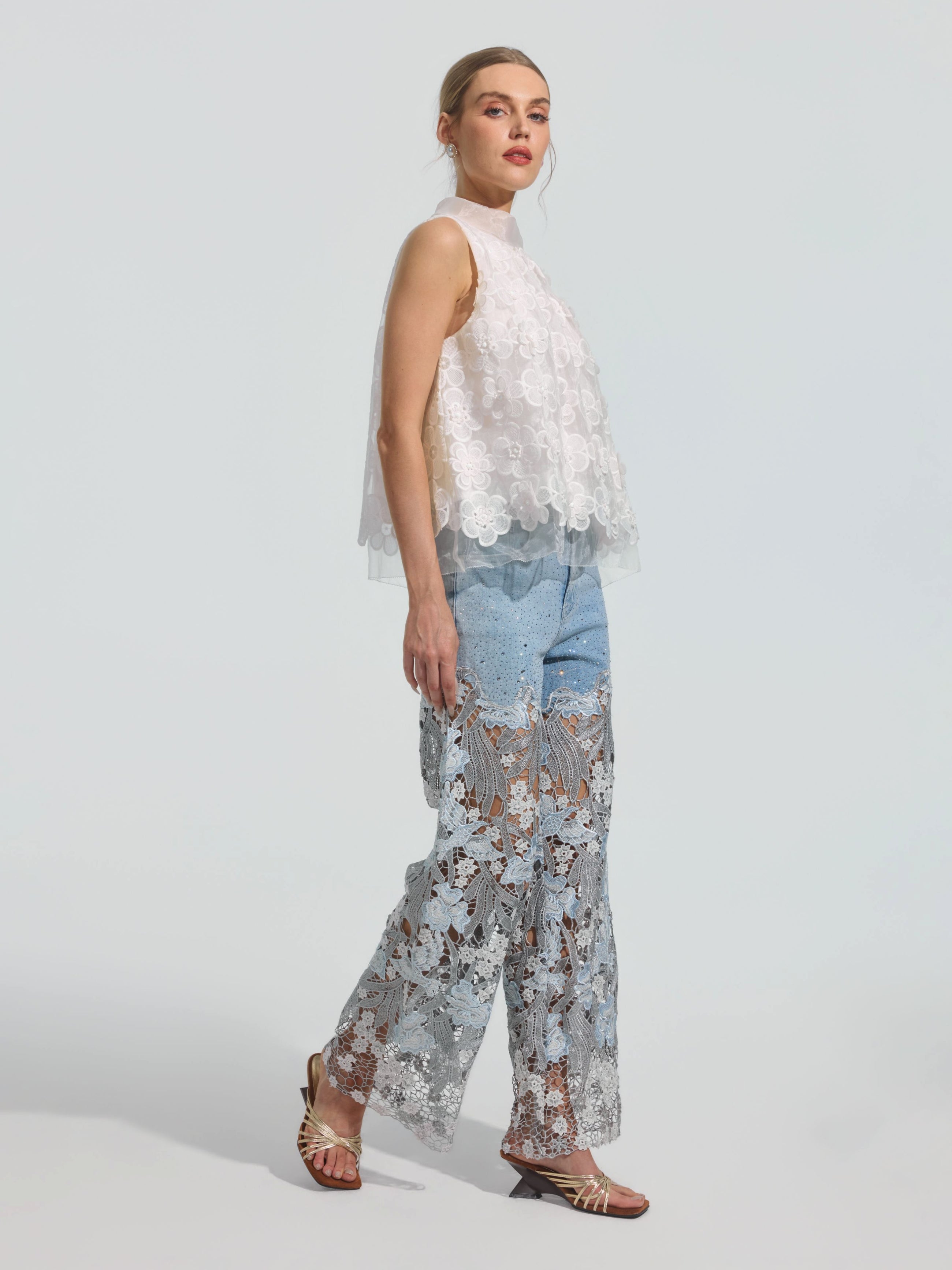 A woman stands in profile against a plain background, wearing the Social Queen 3D Floral Lace Appliqué Silk Top, paired with sheer lace wide-leg pants layered over light blue jeans and open-toed sandals.
