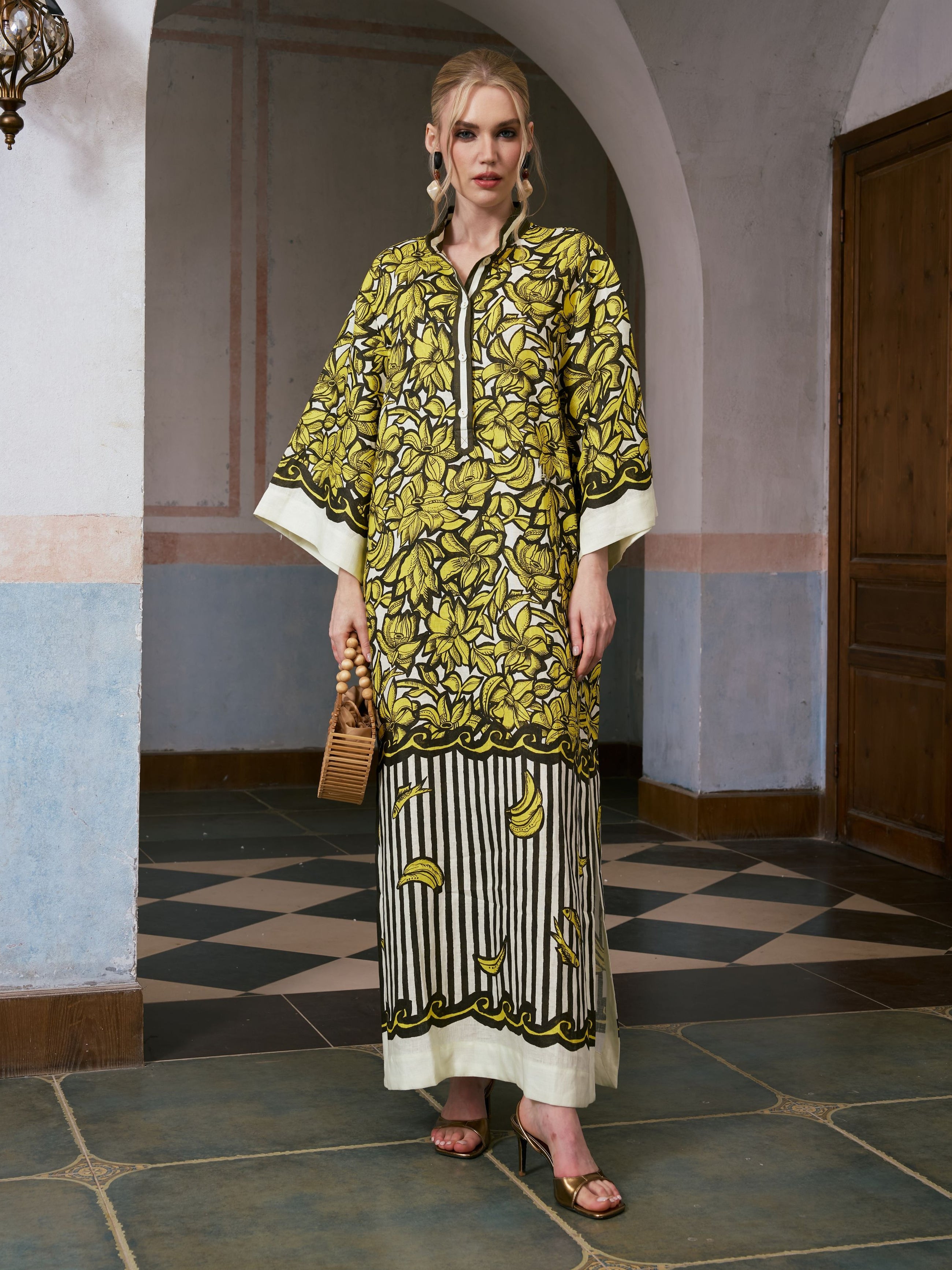 A woman stands indoors wearing the Social Queen Tropical Fruit Print Linen Dress, which features a long, resort wear-inspired silhouette with a vibrant yellow, black, and white tropical fruit pattern. She accessorizes with a wooden handbag and sandals.