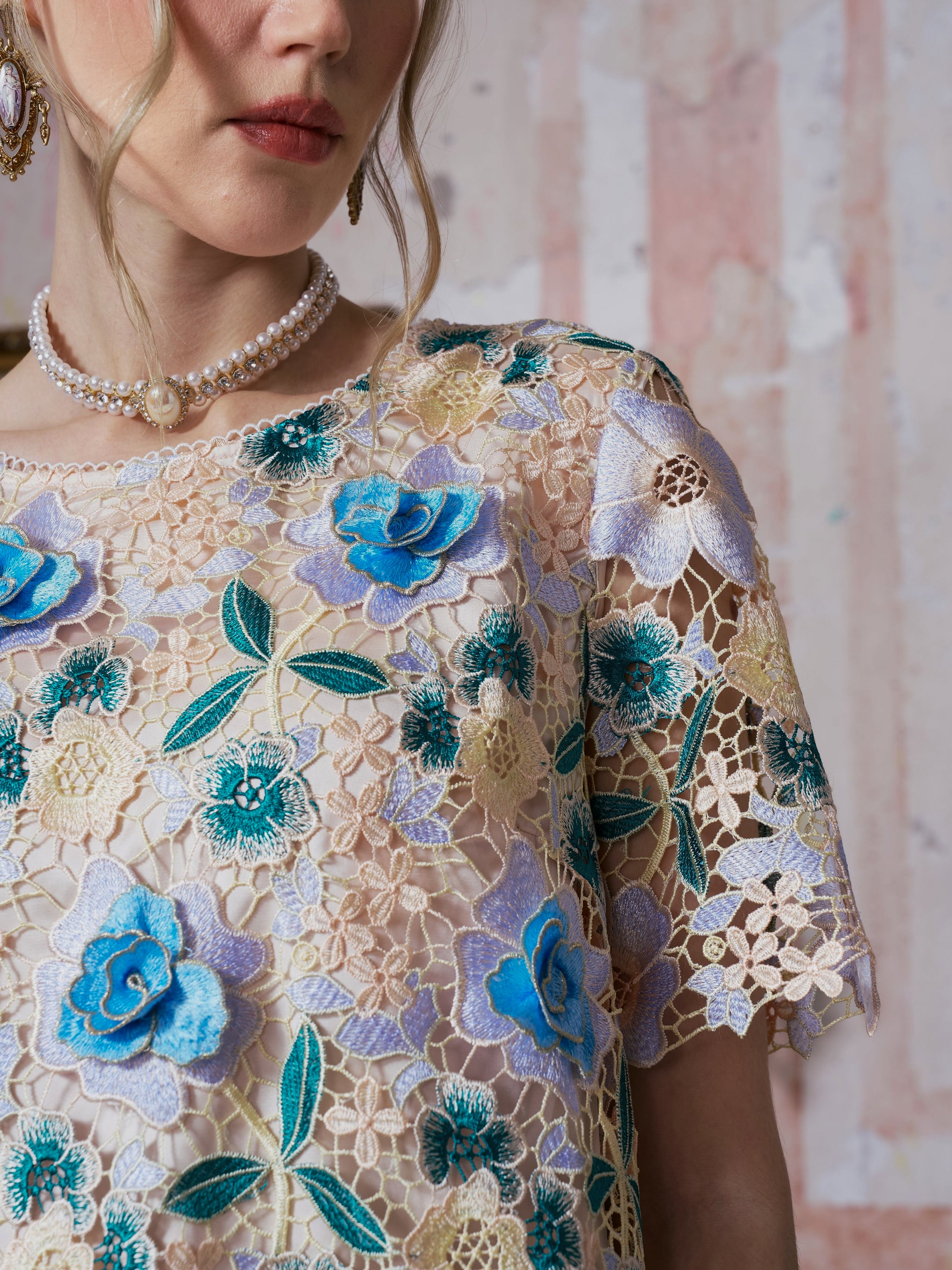 A woman models the Social Queen 3D Floral Cutwork Lace Top, featuring sheer guipure lace with blue and yellow floral embroidery, paired with a pearl necklace and earring; the image is cropped from chin to upper arm against a textured background.