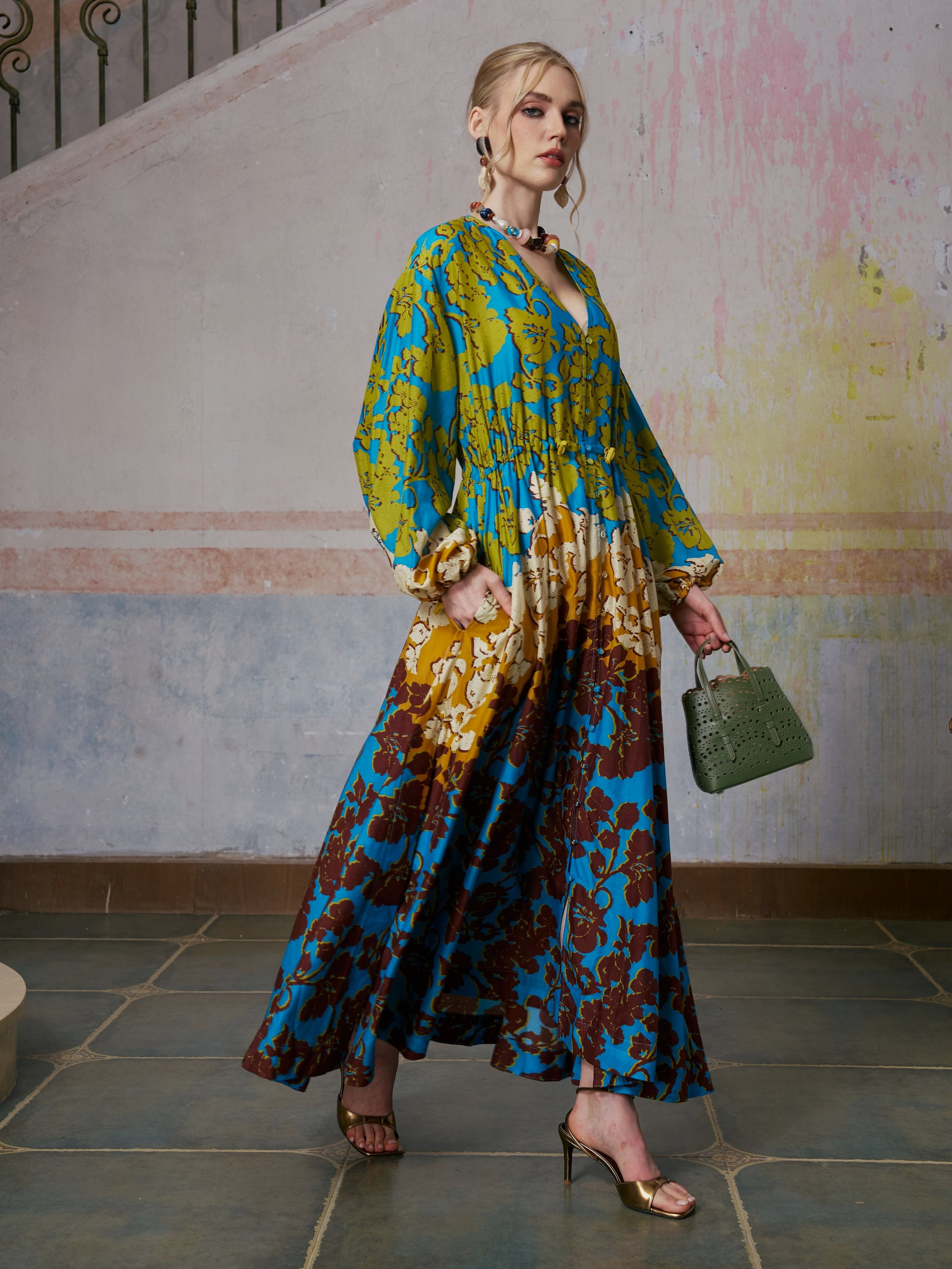 A woman stands indoors against a textured wall, wearing the Social Queen Contrast Floral Print Silk Cotton Dress—a long, colorful piece with blue, yellow, and brown patterns, ideal for chic resort wear—paired with gold high-heeled sandals and a green handbag.