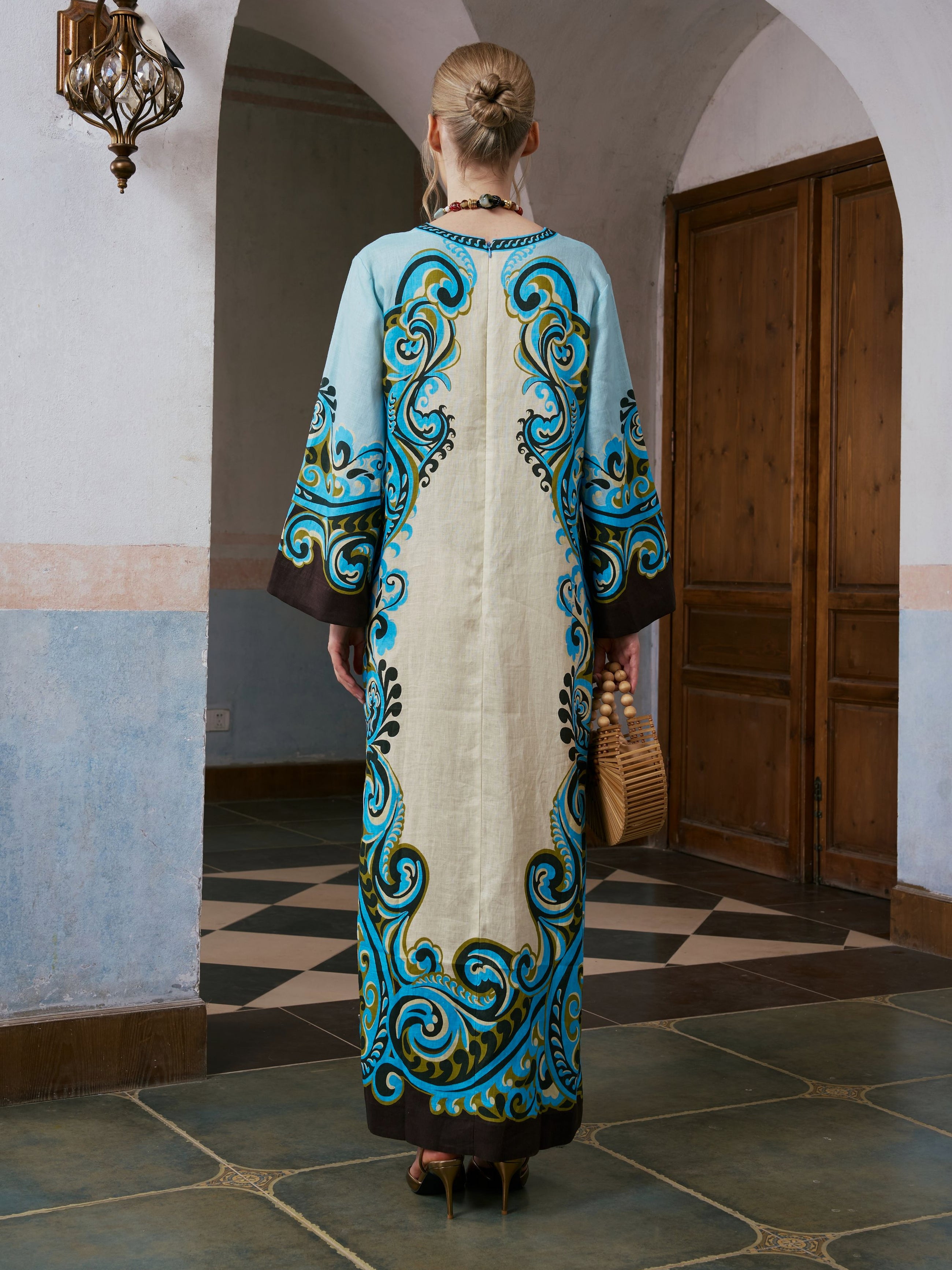A woman stands indoors, facing away, wearing the Paisley Print Linen Dress with blue, black, and gold patterns from Social Queen. She holds a woven handbag and has her hair in a bun.