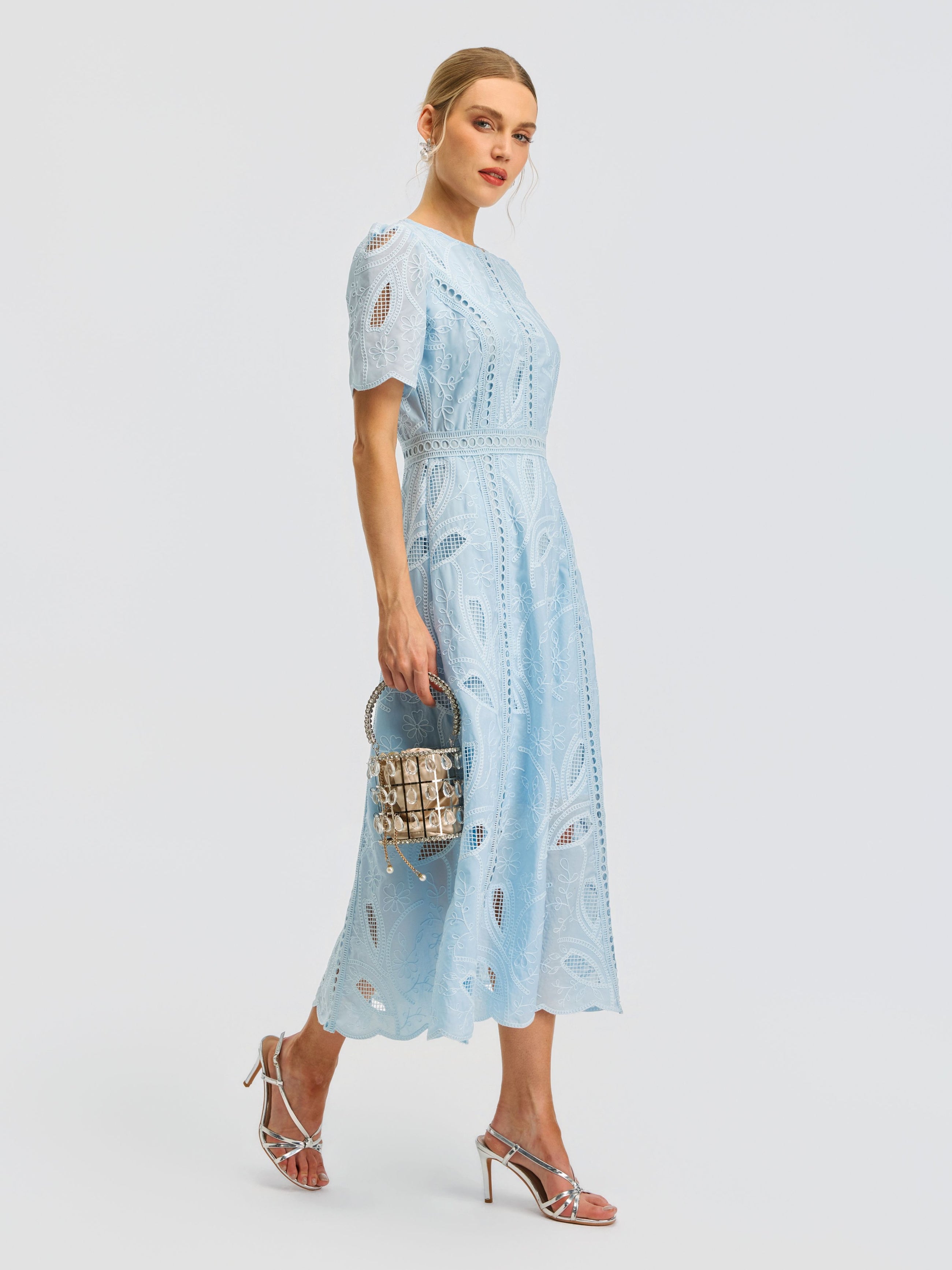 A woman wearing the Social Queen Floral Cutwork Embroidered Dress—a light blue, short-sleeved lace midi dress with delicate floral embroidery—paired with silver high-heeled sandals and a small beaded handbag, stands against a plain white background.