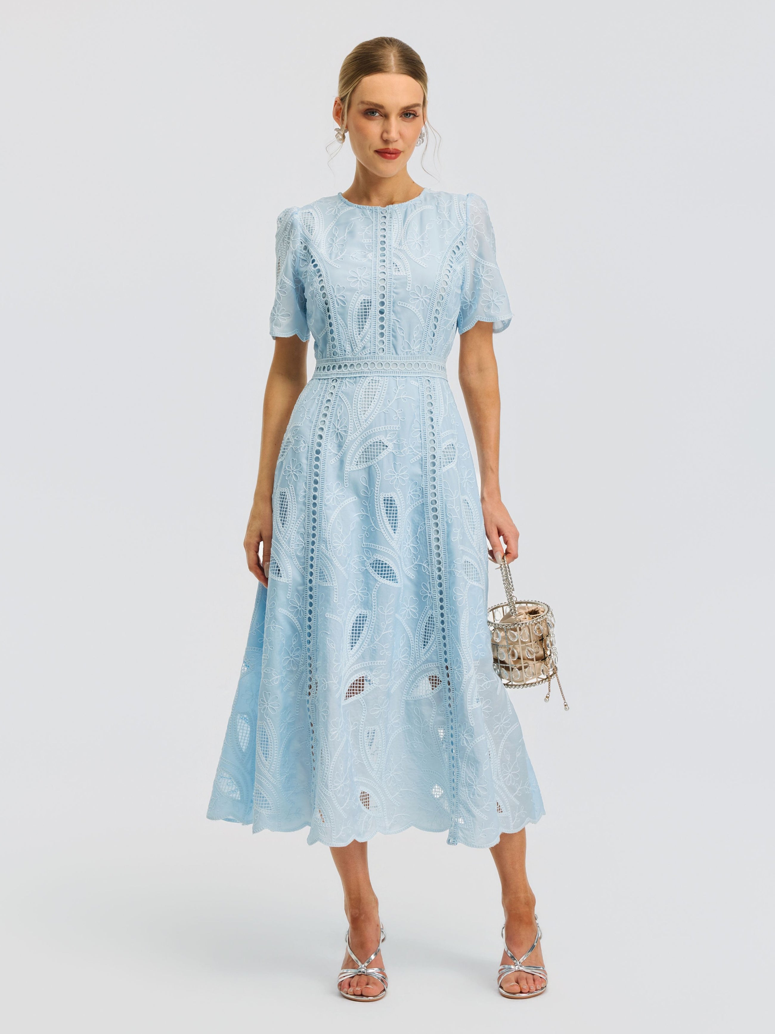 A woman wearing the Social Queen Floral Cutwork Embroidered Dress—a light blue, lace midi dress with short sleeves—holds a small purse while standing against a plain white background.