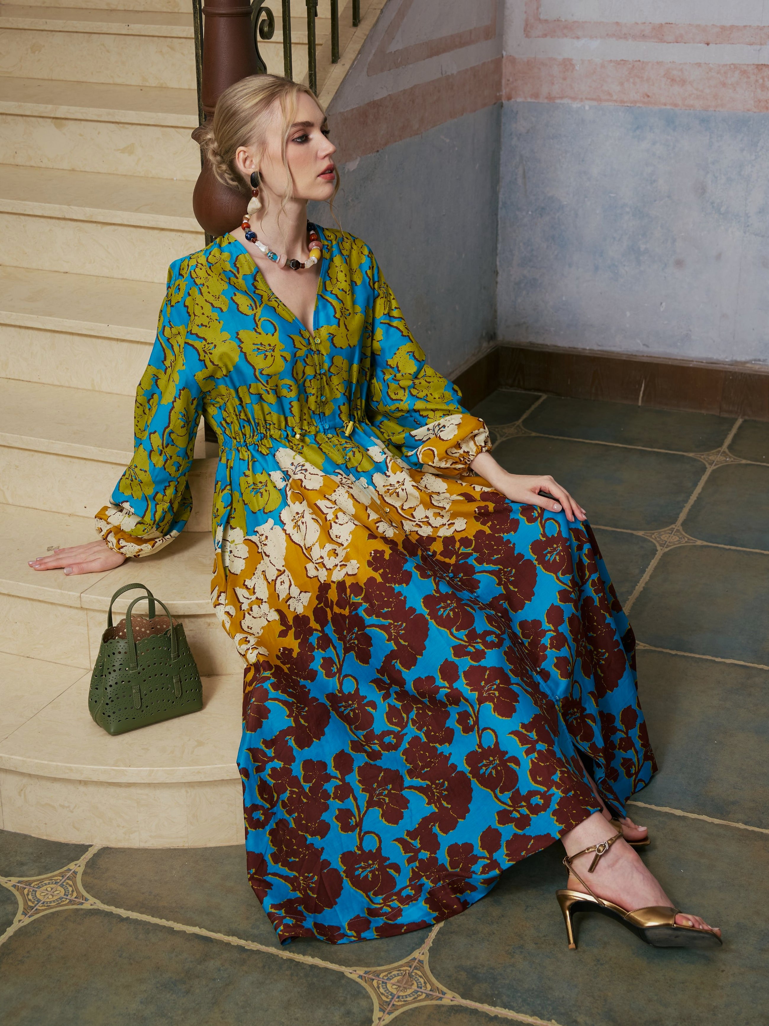 A woman in the Social Queen Contrast Floral Print Silk Cotton Dress sits on tiled steps indoors, a green handbag by her side and one hand resting on the stair. Her silk cotton maxi dress exudes effortless resort wear elegance, lending her a look that is both relaxed and refined.