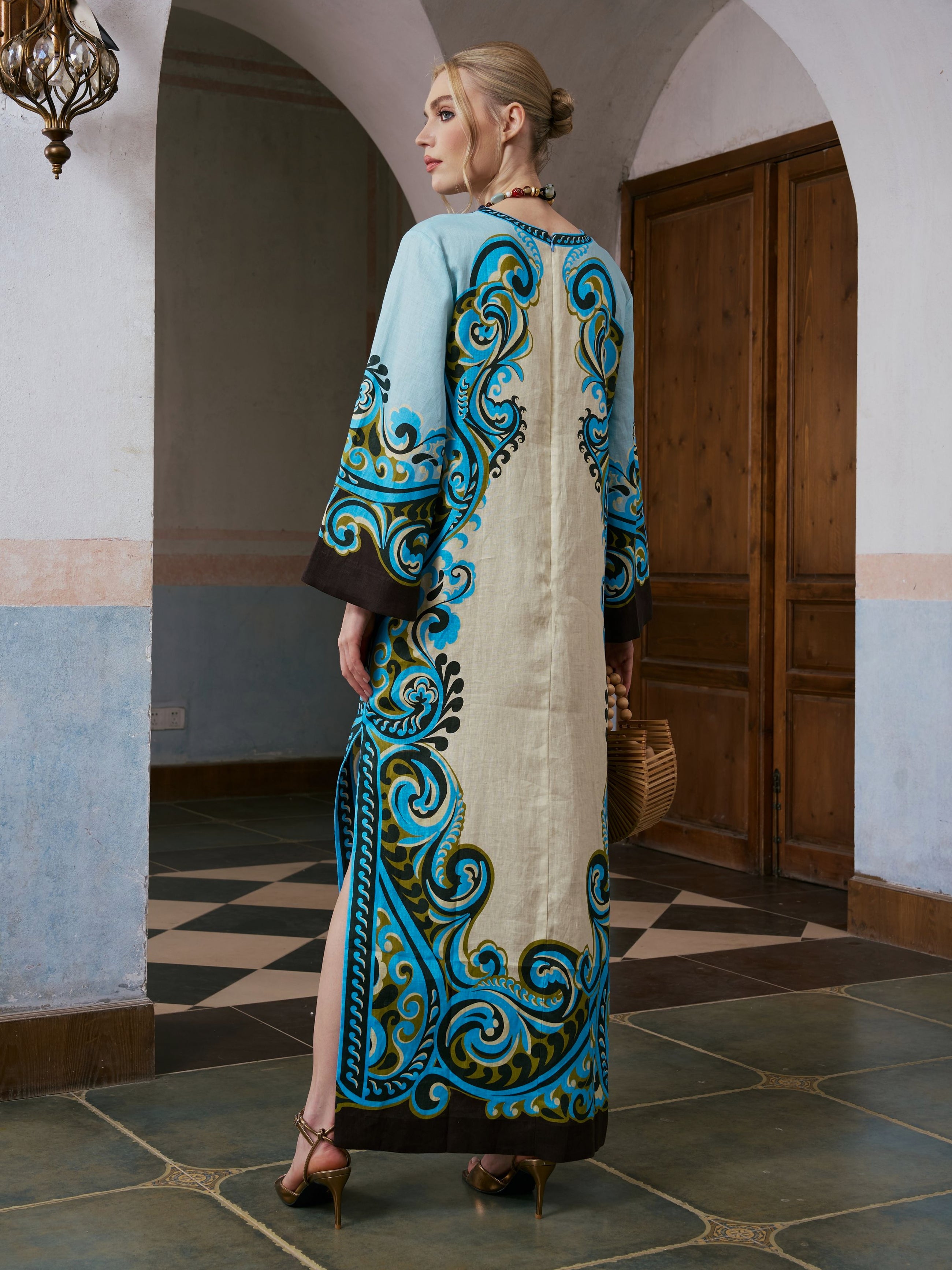 A woman in heels stands indoors, seen from the back, wearing the Social Queen Paisley Print Linen Dress with blue and teal patterns, a paisley border print, and an included belt.