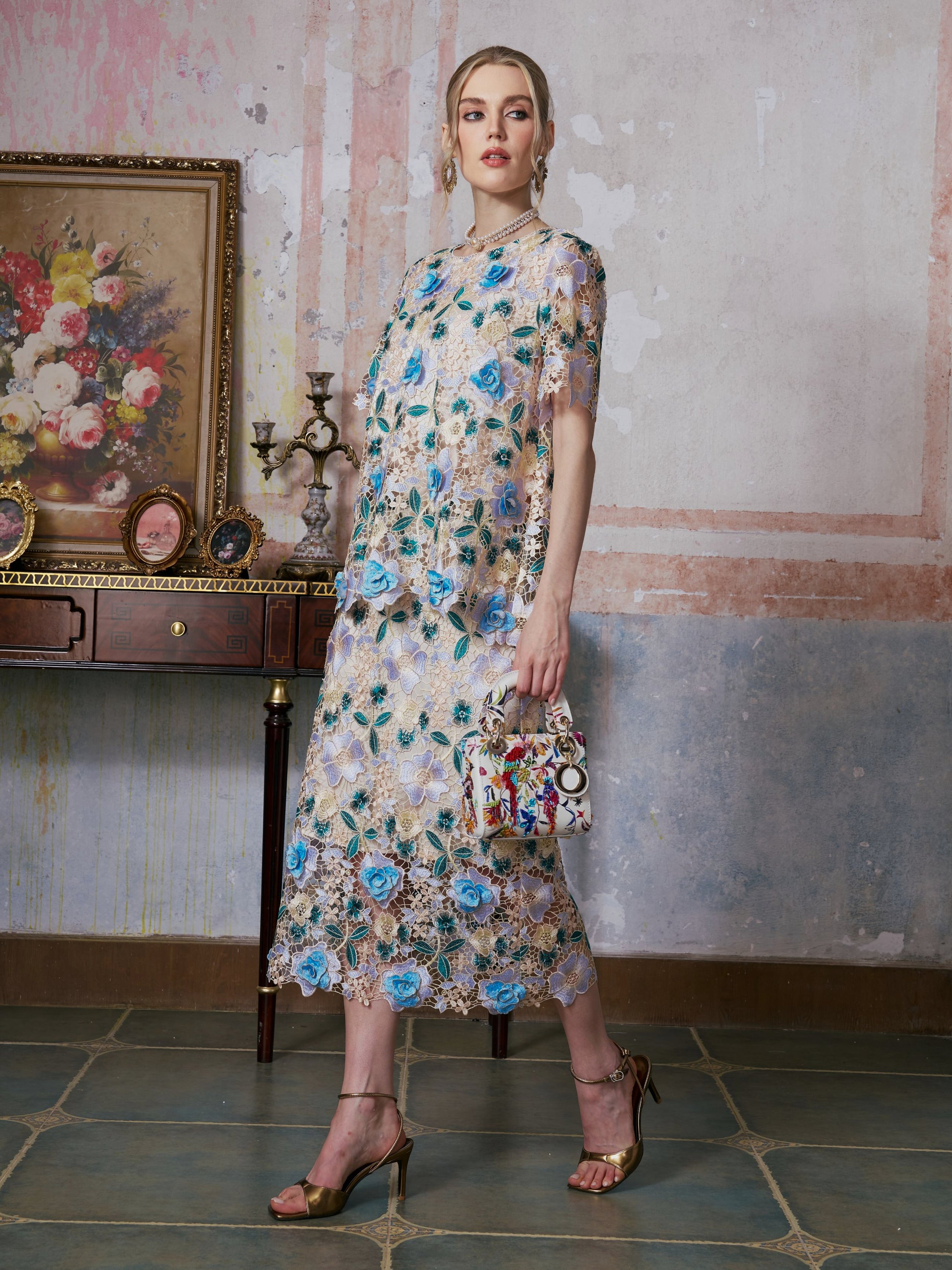 A woman wearing the Social Queen 3D Floral Cutwork Lace Top, adorned with blue flowers and delicate 3D floral appliqués, holds a patterned handbag as she stands in a decorated room featuring vintage furniture and a floral painting.