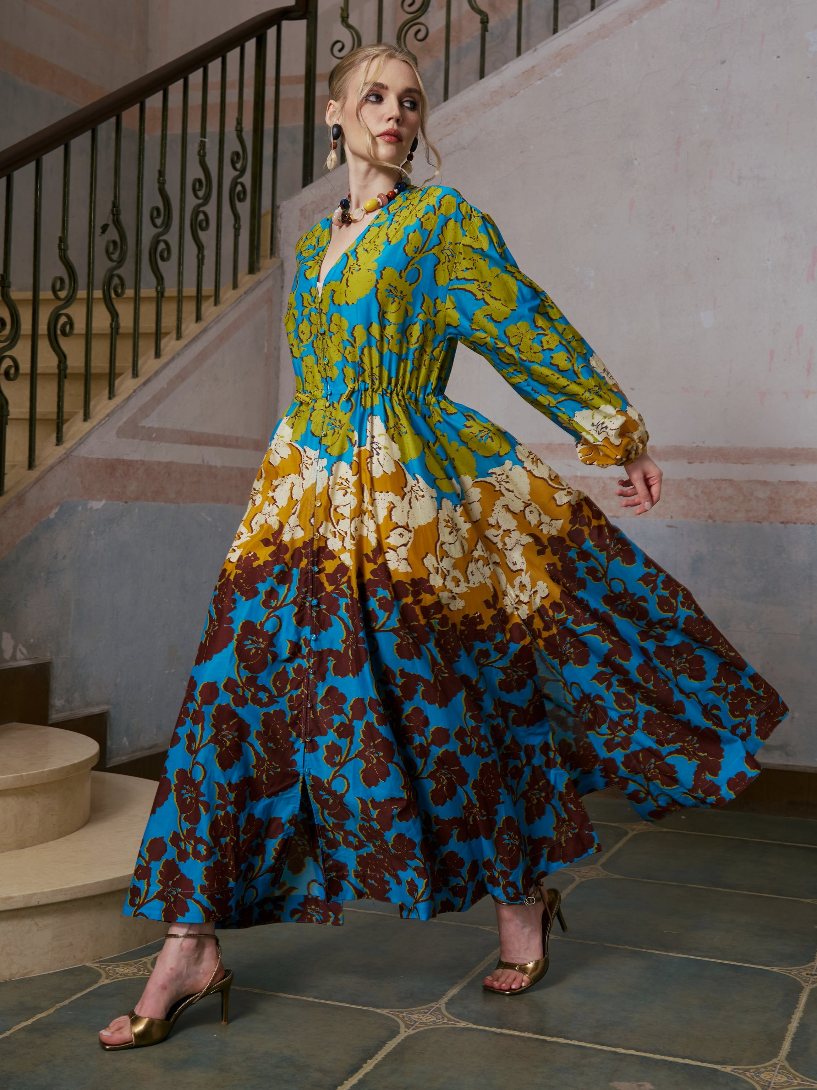 A woman stands indoors near a staircase on a tiled floor, exuding effortless resort wear elegance in the Social Queen Contrast Floral Print Silk Cotton Dress, which features a floral pattern in blue, yellow, and brown tones. She completes the look with gold high-heeled sandals.