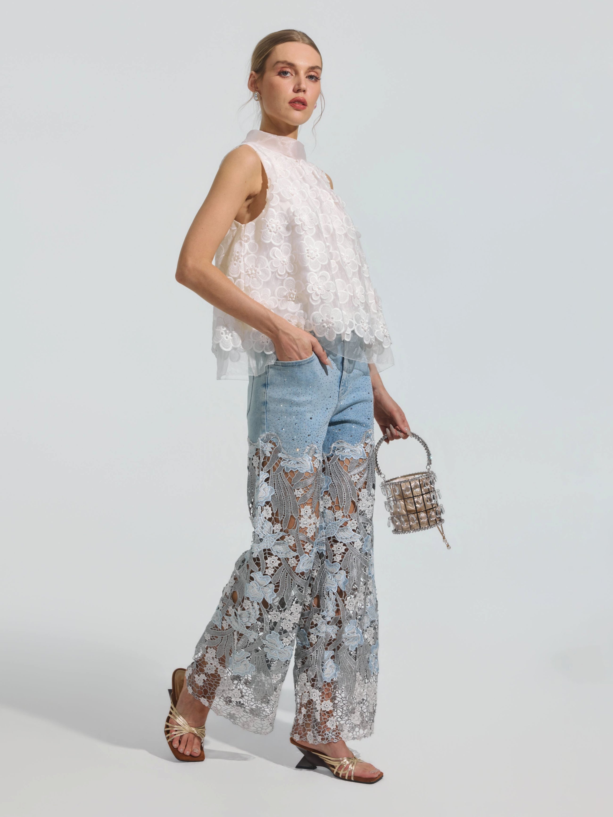 A woman wears the Social Queen Floral Lace Patchwork Pants—a light blue, stretch denim wide-leg style featuring lace and sheer floral patchwork accents—paired with a sleeveless white lace top, flat sandals, and a small bag, posed against a plain background.
