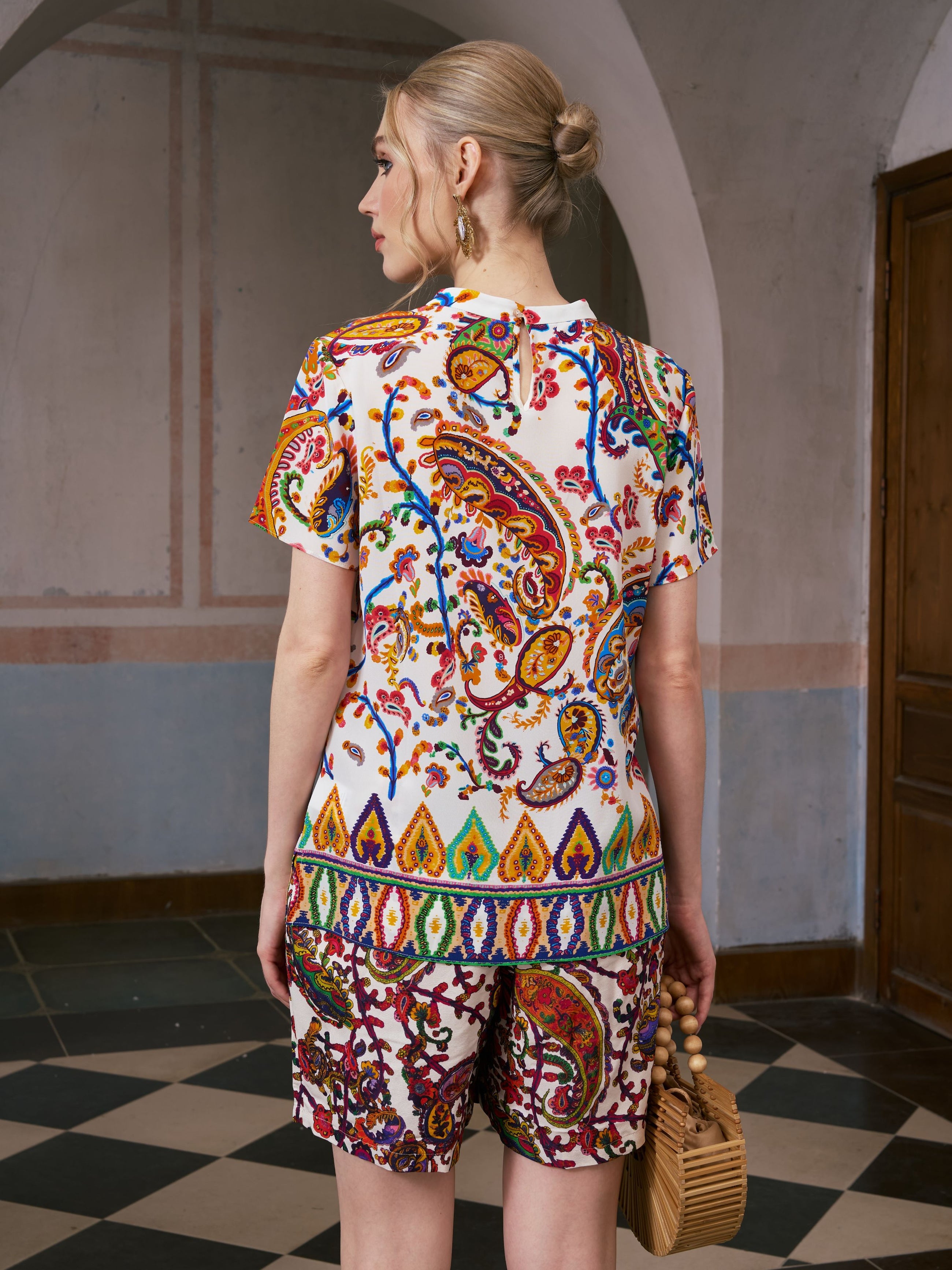 A woman stands indoors with her back to the camera, wearing the Paisley Garden Print Silk Top by Social Queen—perfect for resort wear—and holding a wooden handbag.
