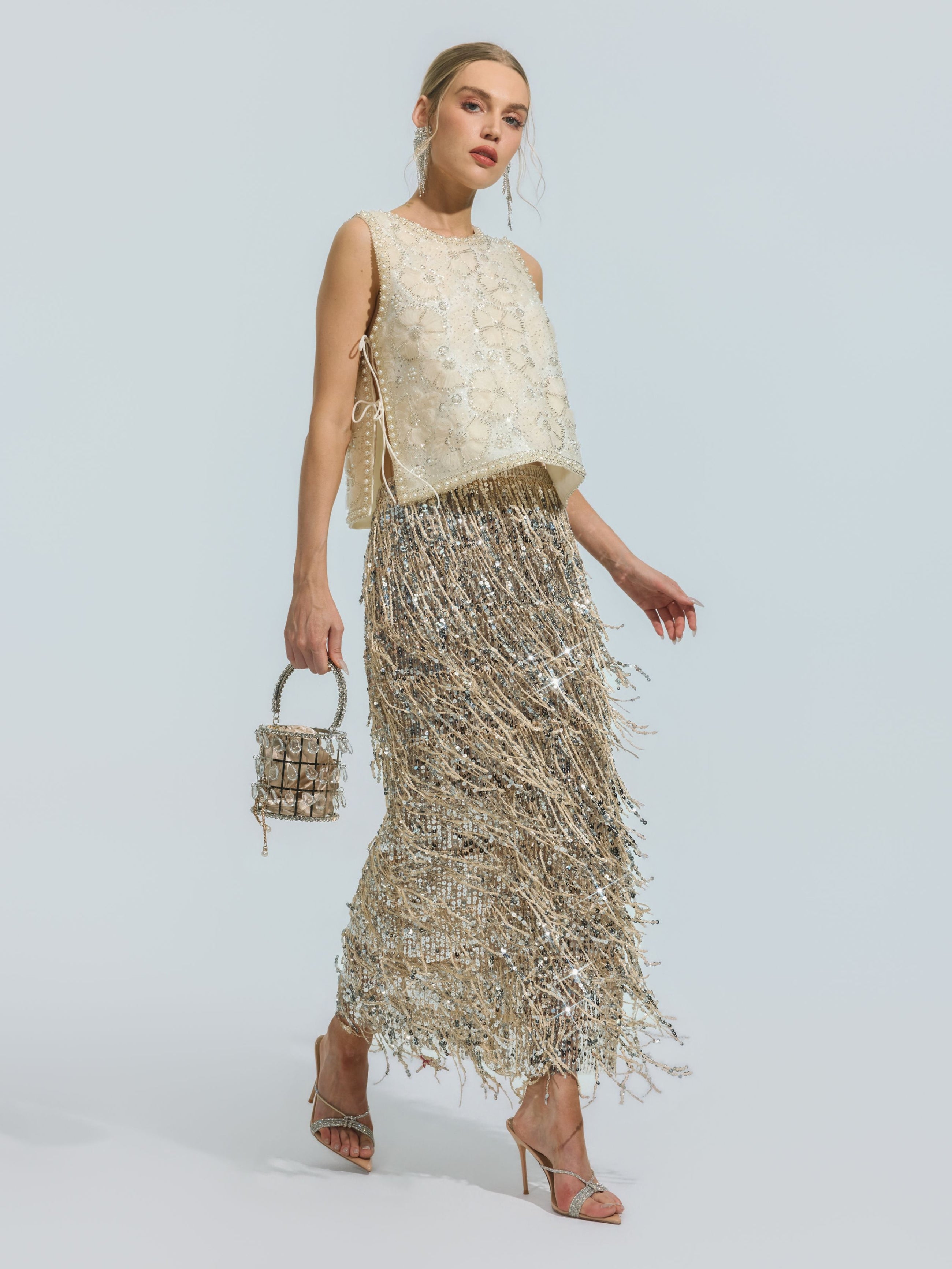 A woman wears the Social Queen Floral Beaded Mesh Sleeveless Top paired with a metallic fringe skirt, accessorized with a small beaded handbag and strappy high heels, set against a plain background.