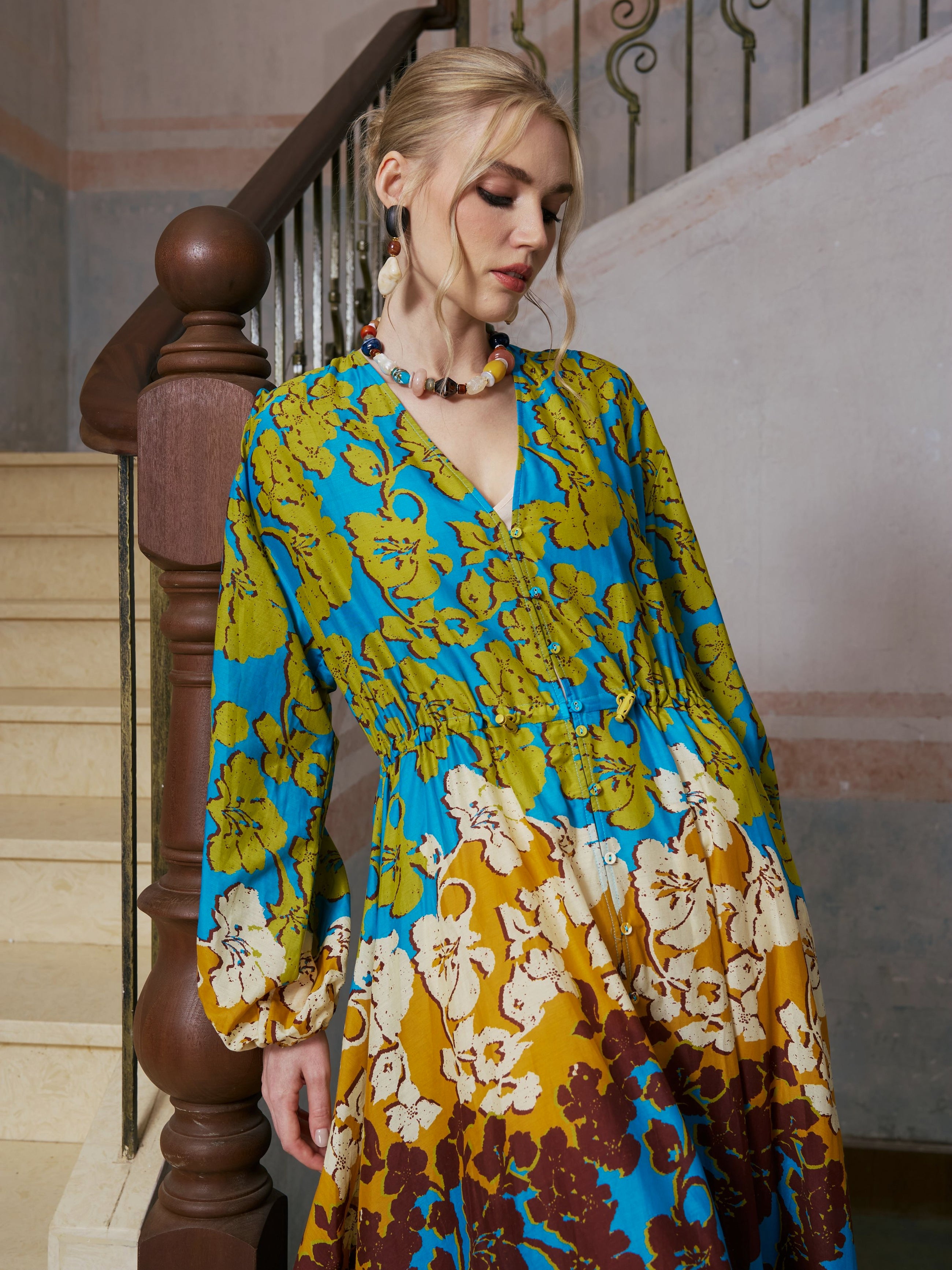 A woman stands on an indoor staircase wearing the Social Queen Contrast Floral Print Silk Cotton Dress, its vivid blue, green, yellow, and brown floral patterns embodying effortless resort style; she gazes downward with a relaxed posture.
