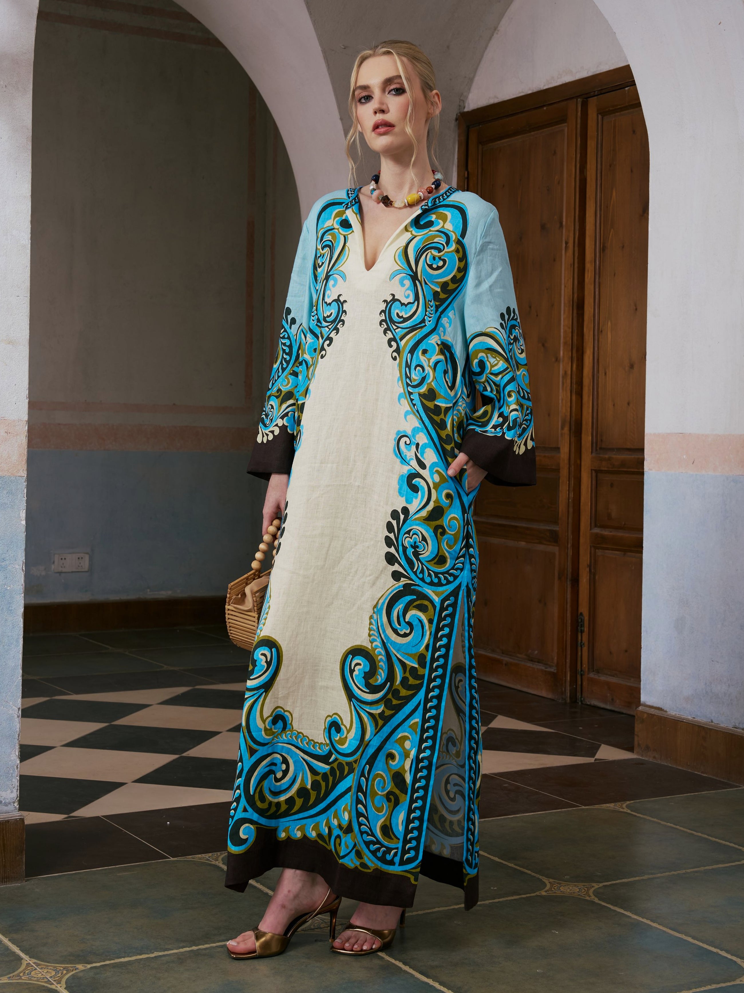 A woman stands indoors wearing the Social Queen Paisley Print Linen Dress with its cream and turquoise paisley border print, black details, and included belt—an elegant linen maxi—paired with a small woven bag and heeled sandals.