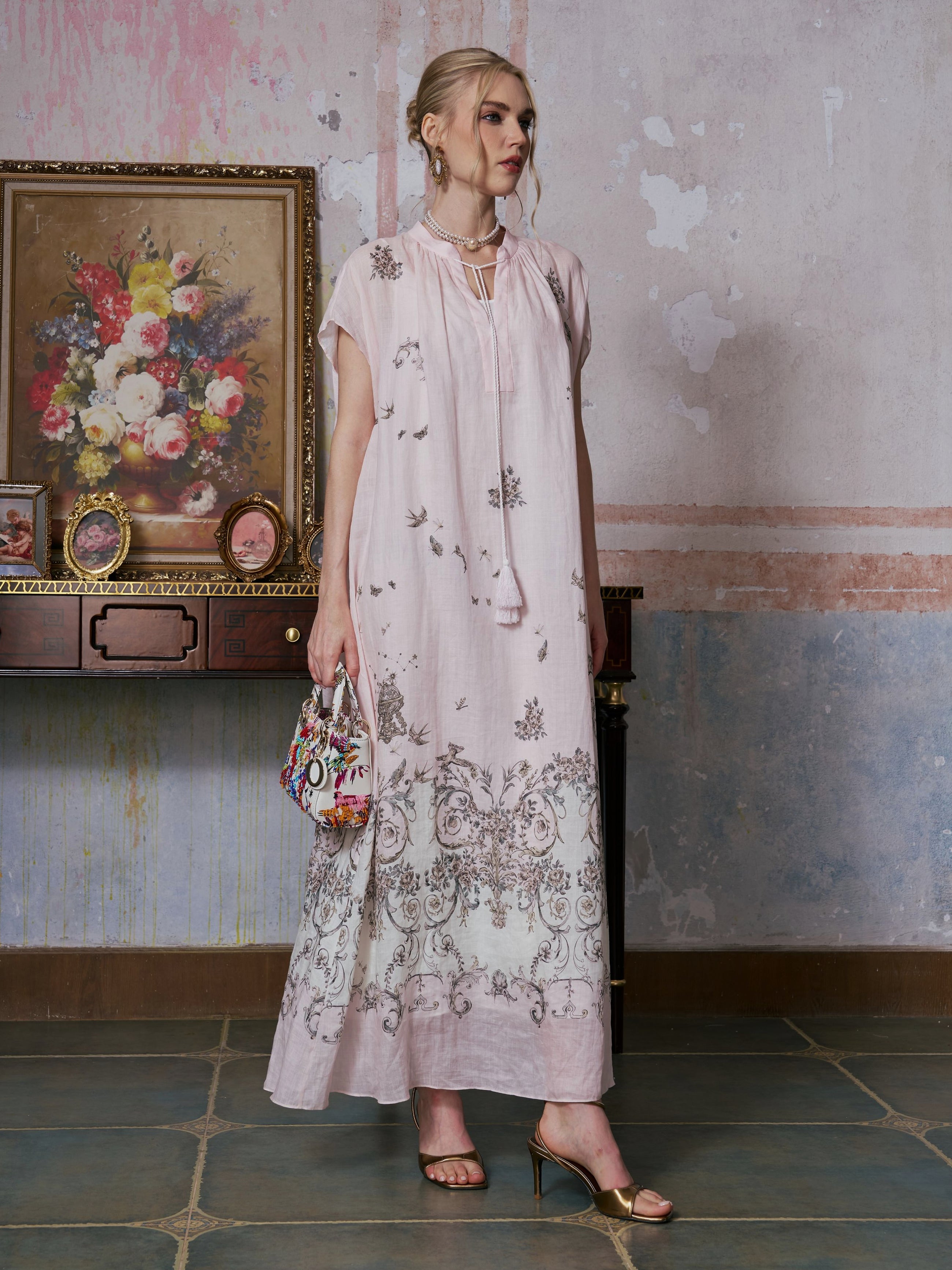 A woman wearing the Social Queen Baroque Garden Print Ramie Dress (includes slip dress), featuring a pale pink, vintage-inspired design with delicate floral embroidery, stands indoors beside a vintage desk and a framed floral painting, holding a colorful handbag.