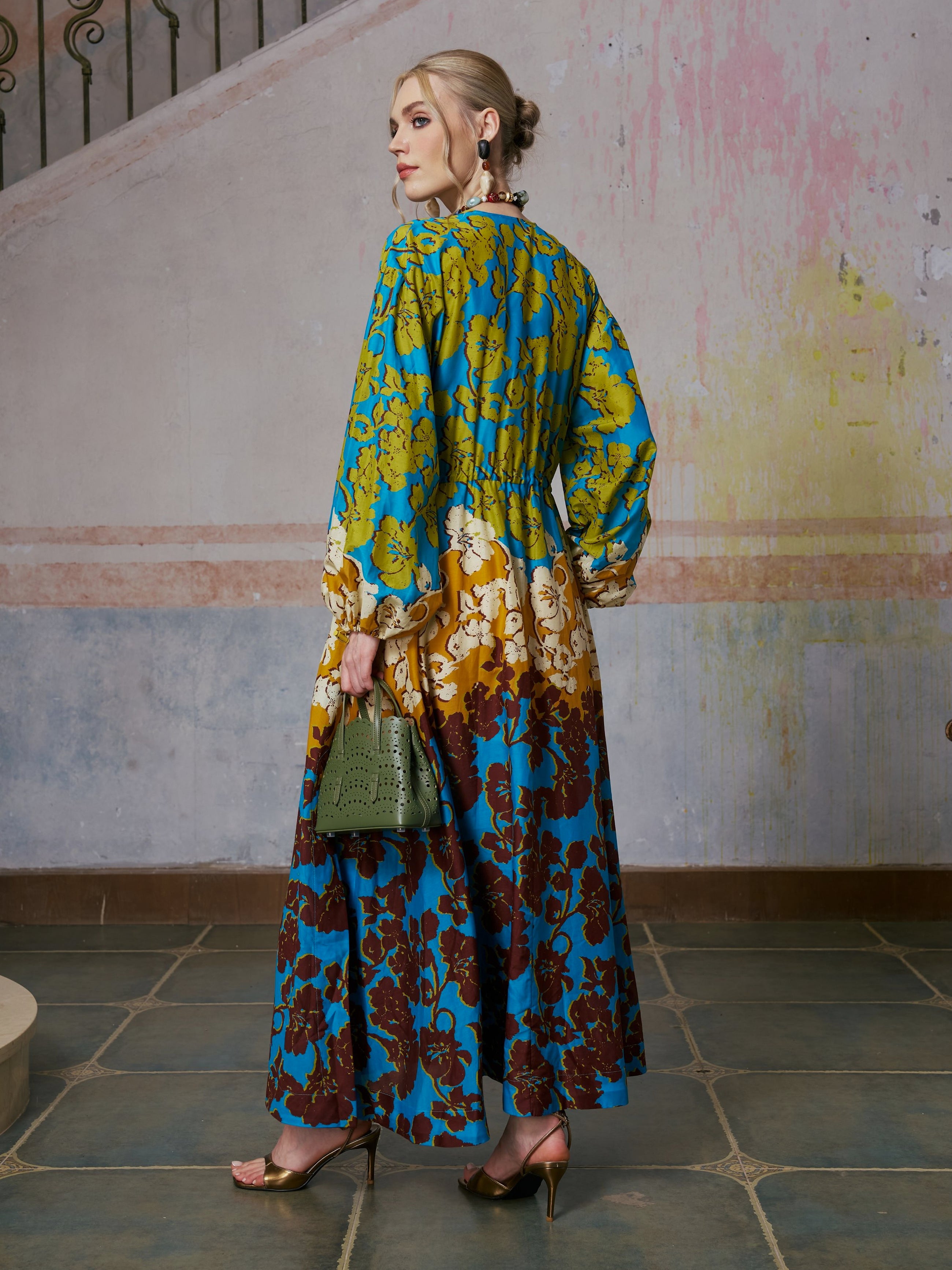 A woman in the Social Queen Contrast Floral Print Silk Cotton Dress and high heels stands on tiled flooring, holding a green bag and looking over her shoulder towards the camera—a perfect example of chic resort wear.