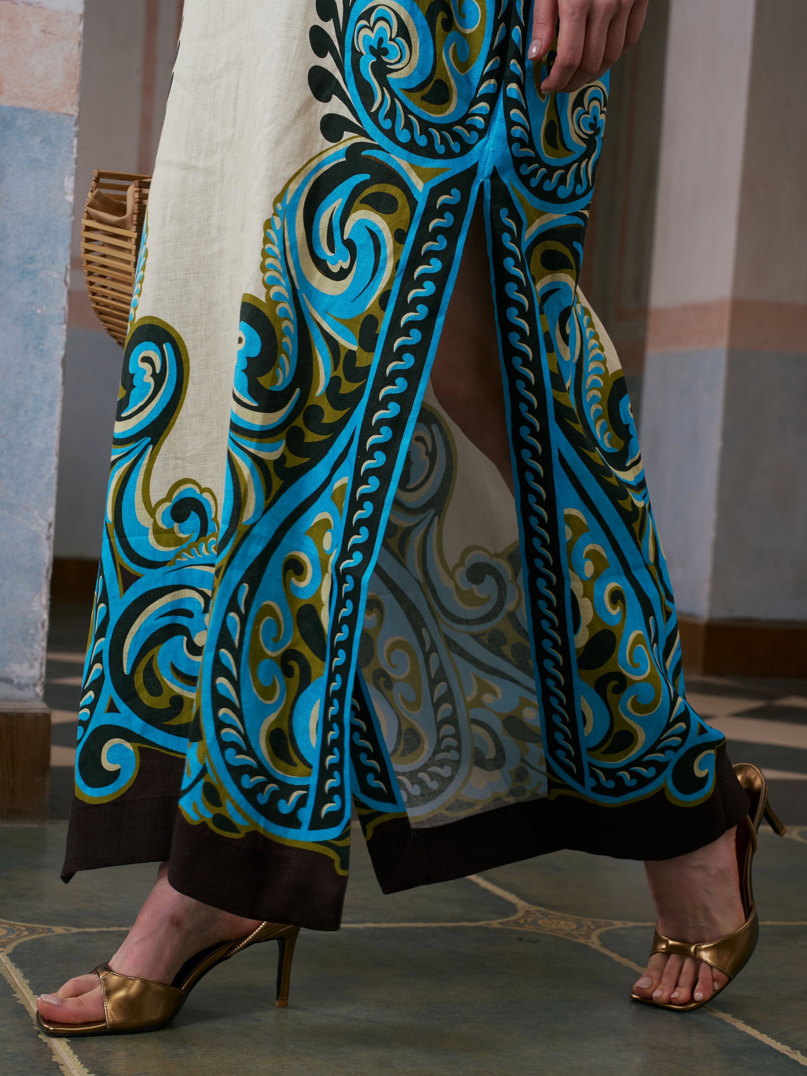 A person wearing the Social Queen Paisley Print Linen Dress (belt included), featuring blue, green, and black designs, pairs it with gold high-heeled sandals while standing indoors on a tiled floor.