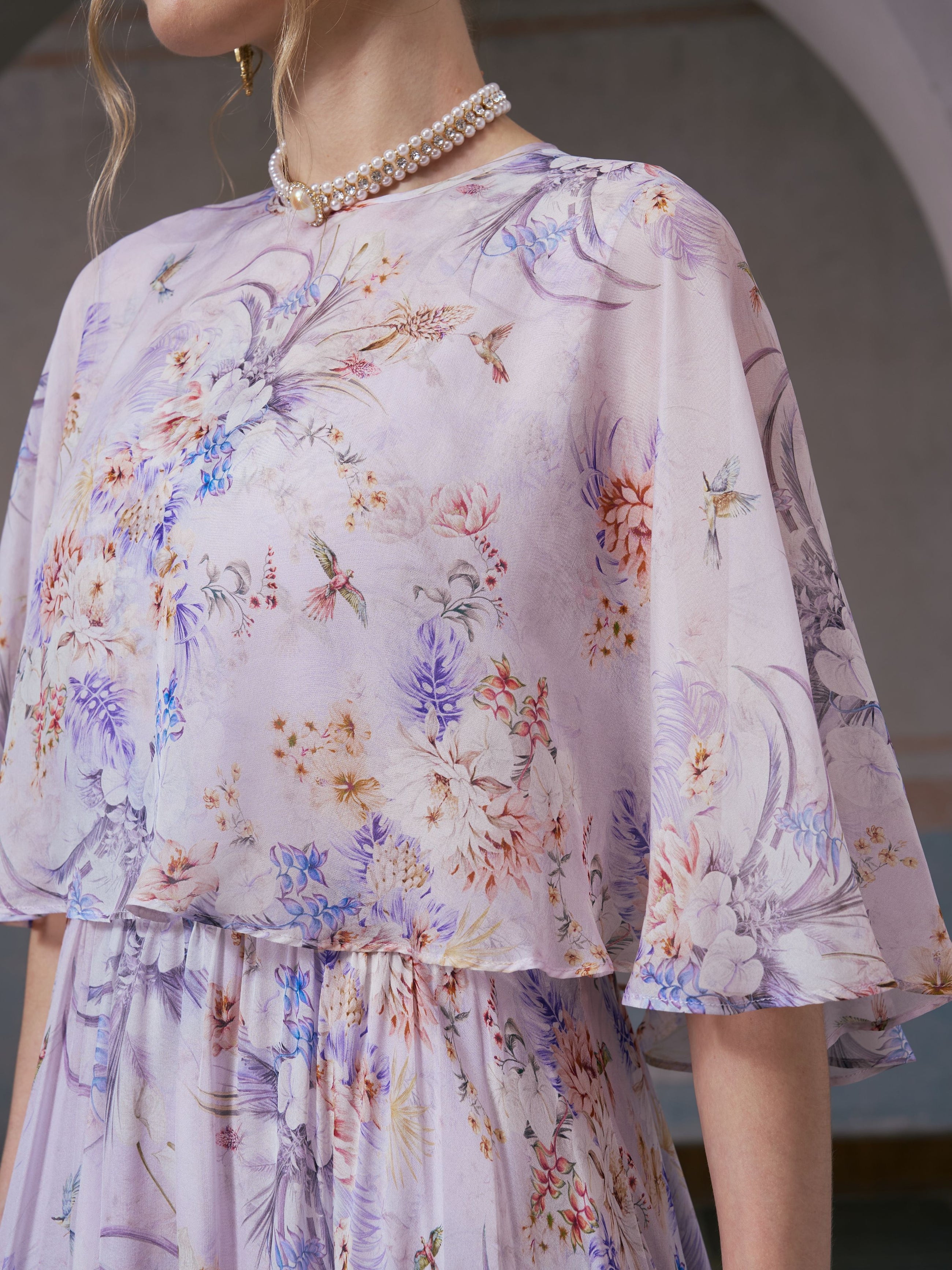 A woman wearing the Social Queen Lavender Print Silk Dress, featuring wide sleeves and a delicate lavender bird motif, paired with a pearl necklace, stands indoors.