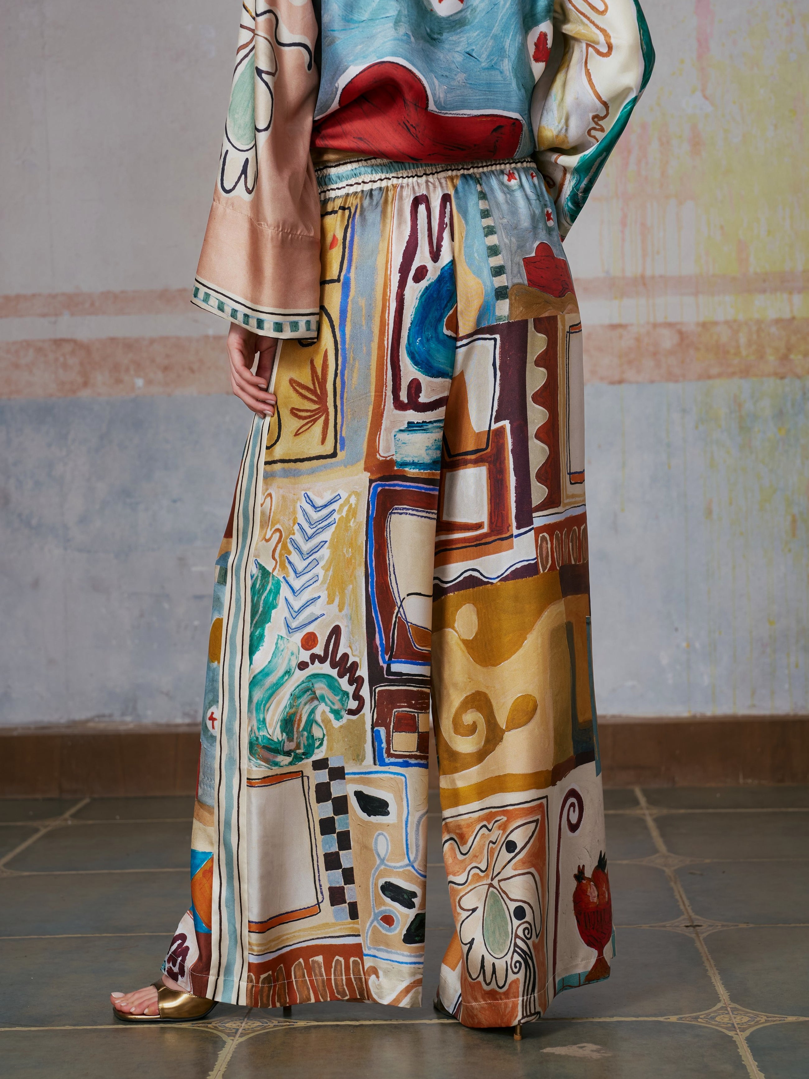 A person wearing Social Queen's Abstract Print Mulberry Silk Pants stands barefoot on a tiled floor against a painted wall.