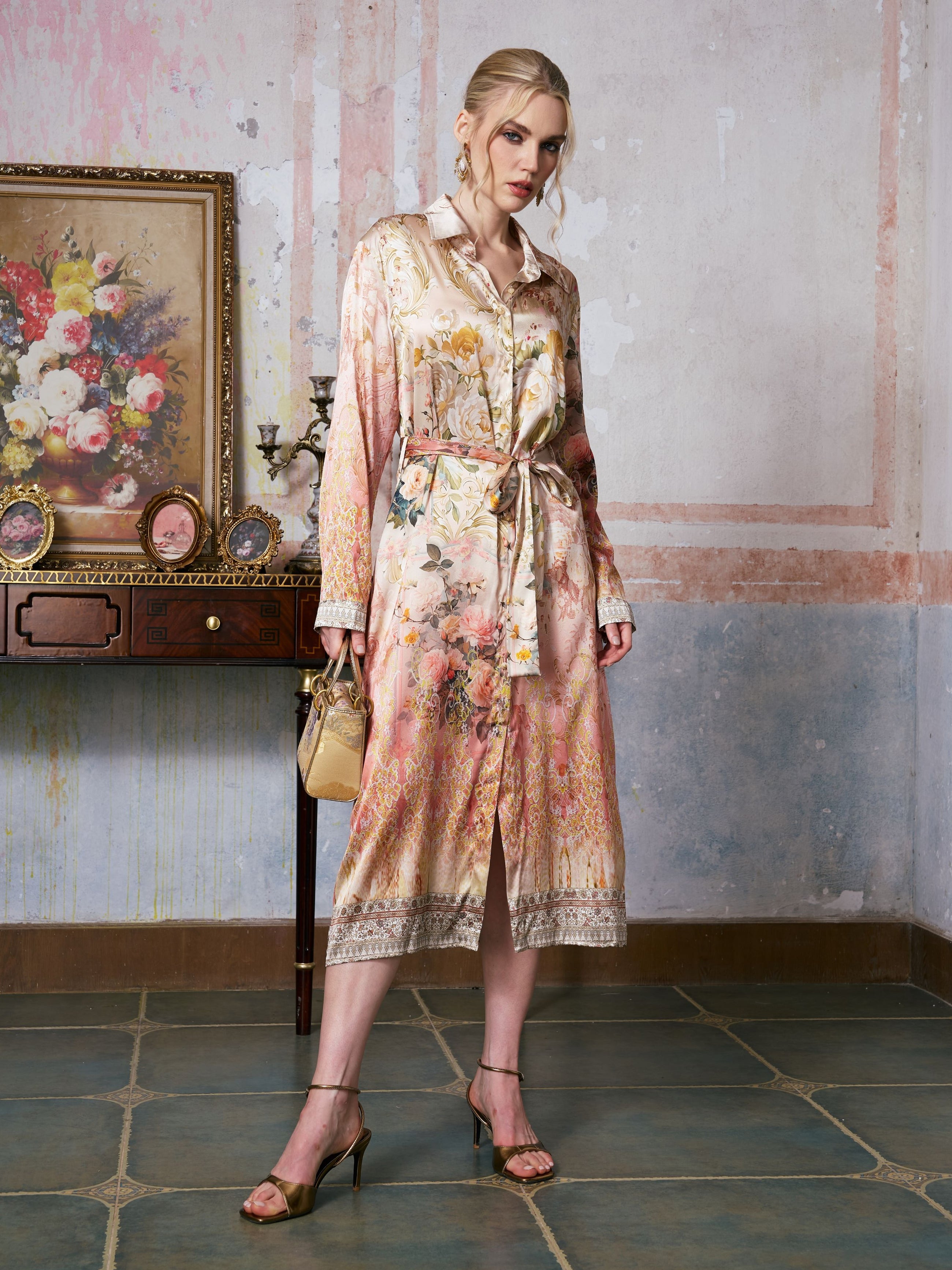 A woman stands indoors wearing the Social Queen Garden Print Mulberry Silk Shirt Dress, belted at the waist and holding a small handbag, with a vintage desk and framed floral painting in the background.