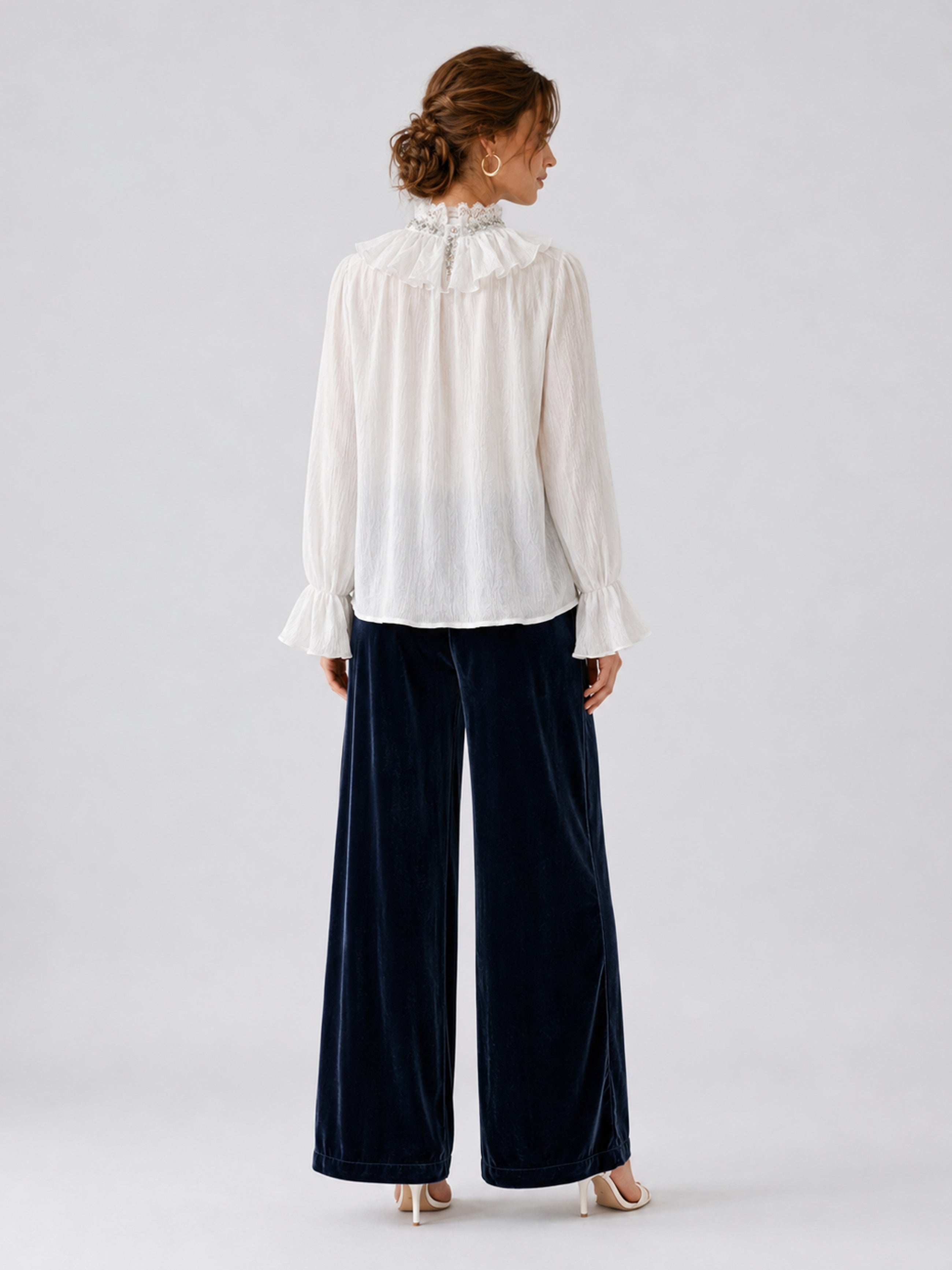 A woman stands facing away, wearing a white long-sleeve blouse with a ruffled collar and cuffs, paired with Social Queen’s Silk Velvet Wide-Leg Pants in navy crinkled texture and white heels, set against a plain light background.