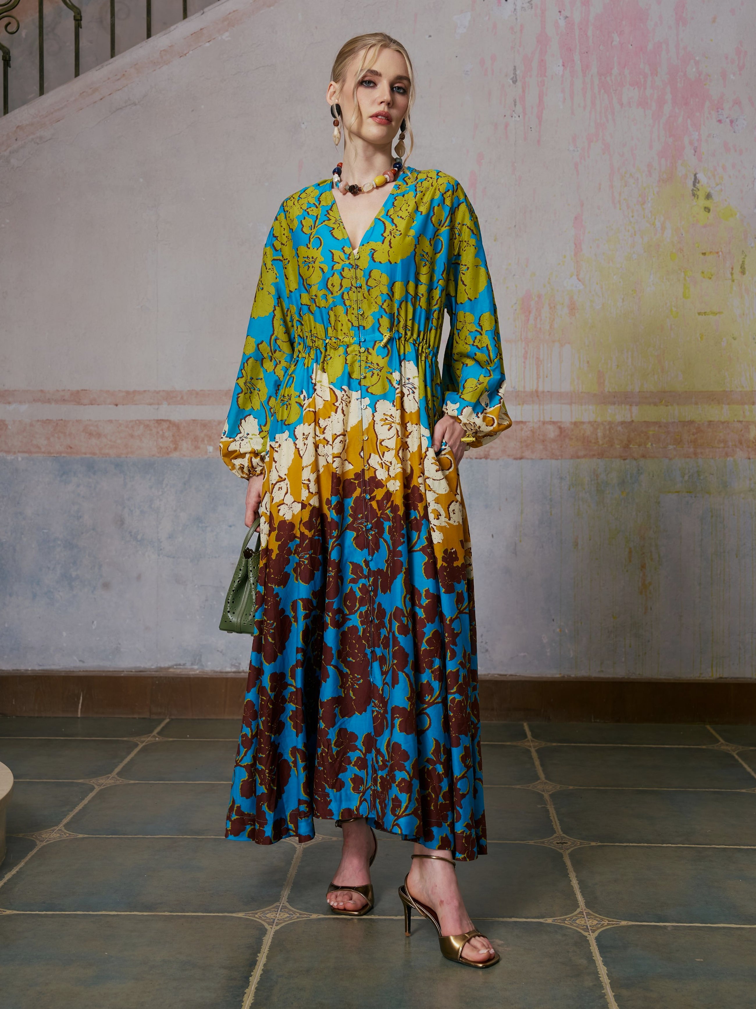 A woman stands indoors wearing the Social Queen Contrast Floral Print Silk Cotton Dress, showcasing a vibrant floral pattern in blue, yellow, and brown tones. She completes the elegant resort wear look with gold heels and jewelry, set against a backdrop of a painted wall and tiled floor.