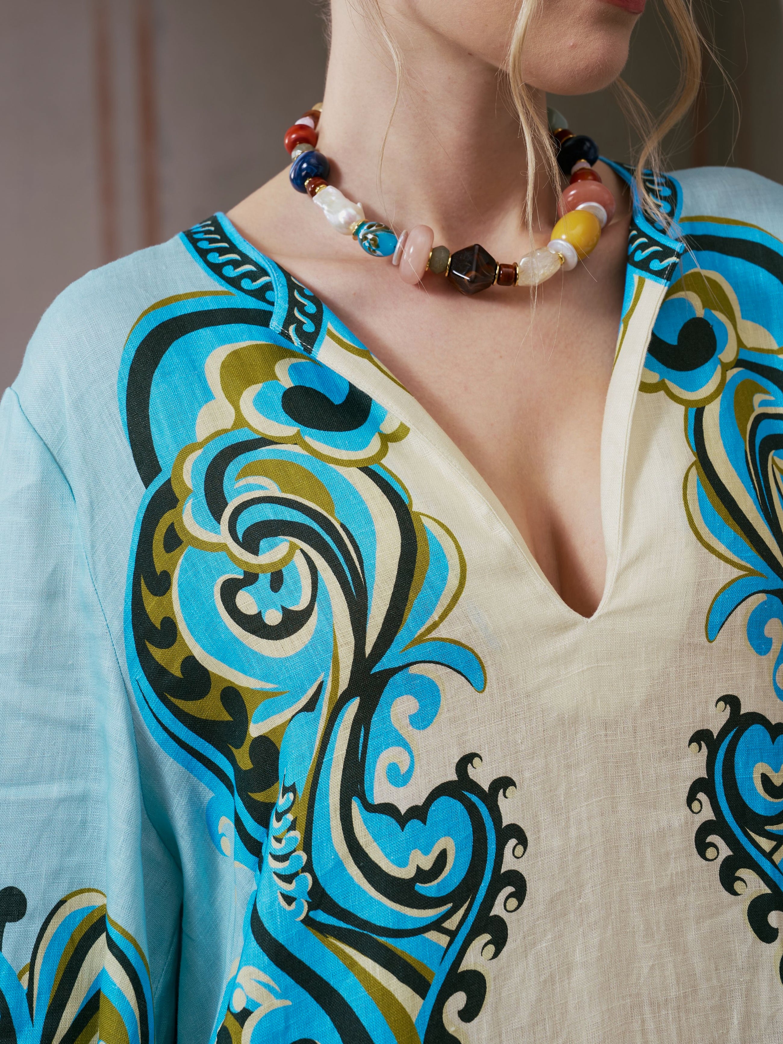 A woman wearing the Social Queen Paisley Print Linen Dress, featuring a vibrant paisley border design and a deep neckline, accessorized with a chunky beaded necklace.