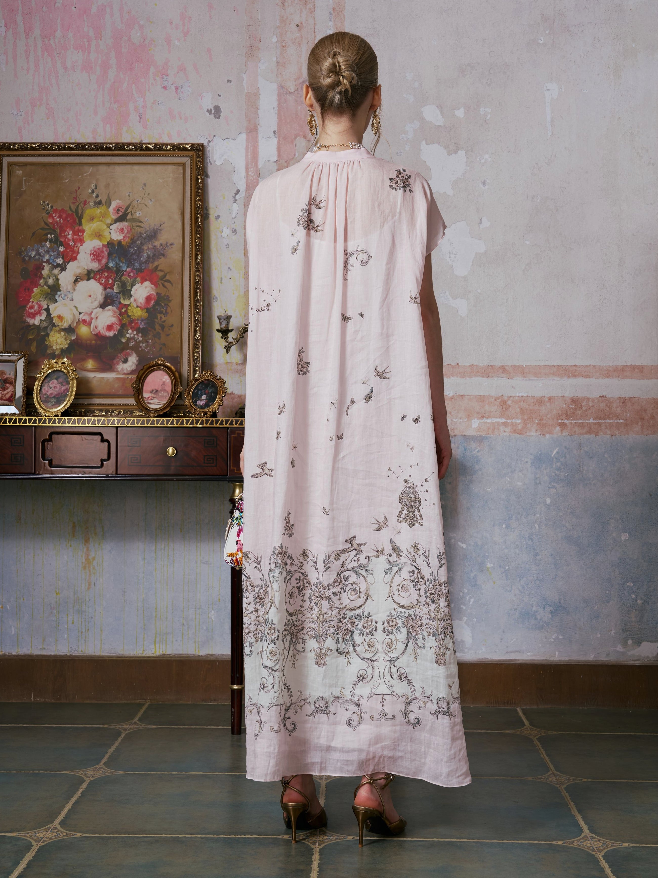 A woman stands facing a wall, wearing the Social Queen Baroque Garden Print Ramie Dress (includes slip dress) in a long, light pink, vintage-inspired style paired with heels. A framed floral painting and small objects rest on a wooden table beside her.