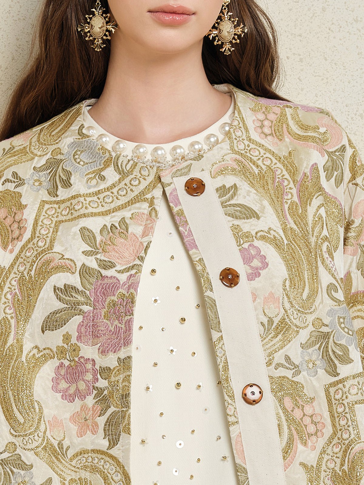 A woman wears the Social Queen Italian Baroque Floral Metallic Jacquard Jacket with brown buttons over a pearl-embellished white top, paired with ornate gold earrings.
