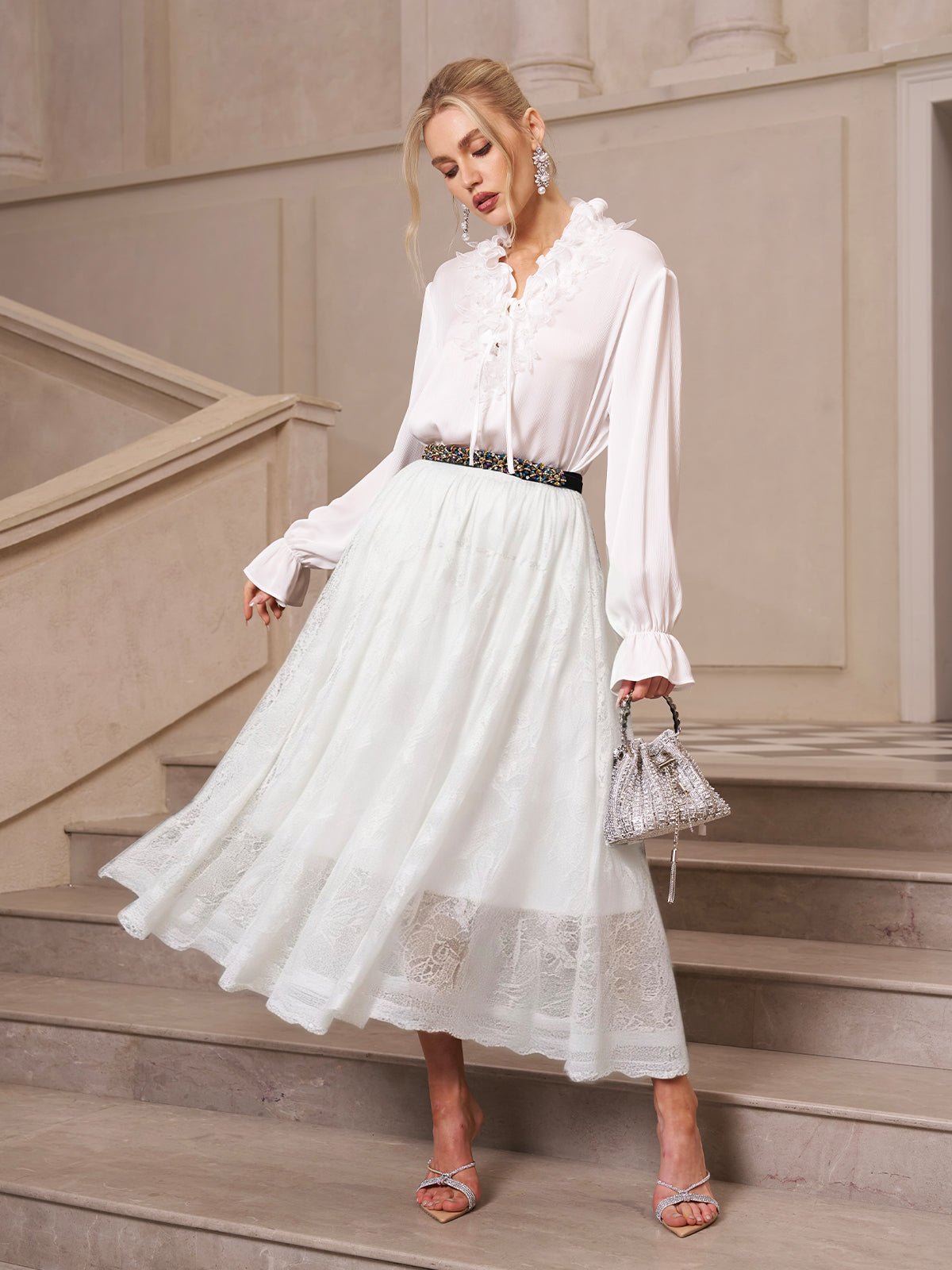 A woman stands on stairs wearing a white ruffled blouse, the Social Queen Jacquard Lace A-Line Midi Skirt, silver heels, and holding a small silver handbag.
