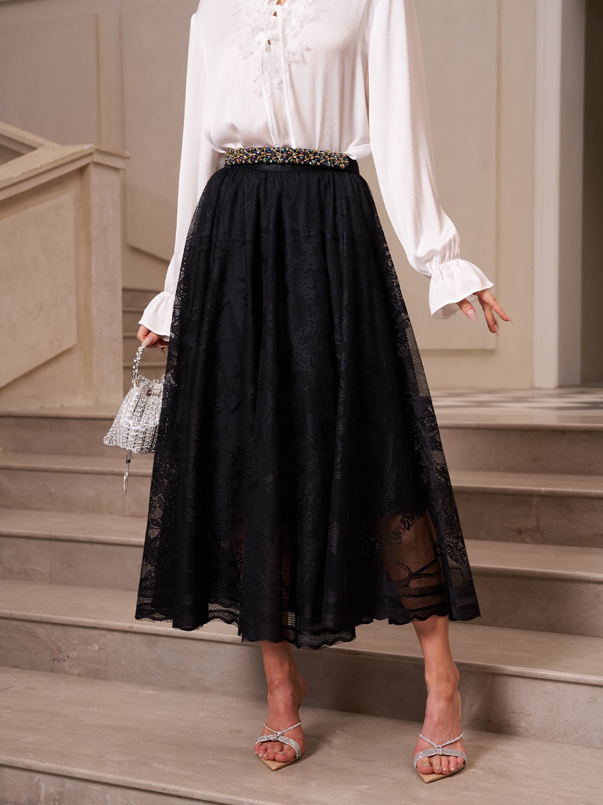 A person wears a white long-sleeve blouse, the Social Queen Jacquard Lace A-Line Midi Skirt with a beaded waistband, silver heeled sandals, and holds a small silver handbag while standing on stairs.