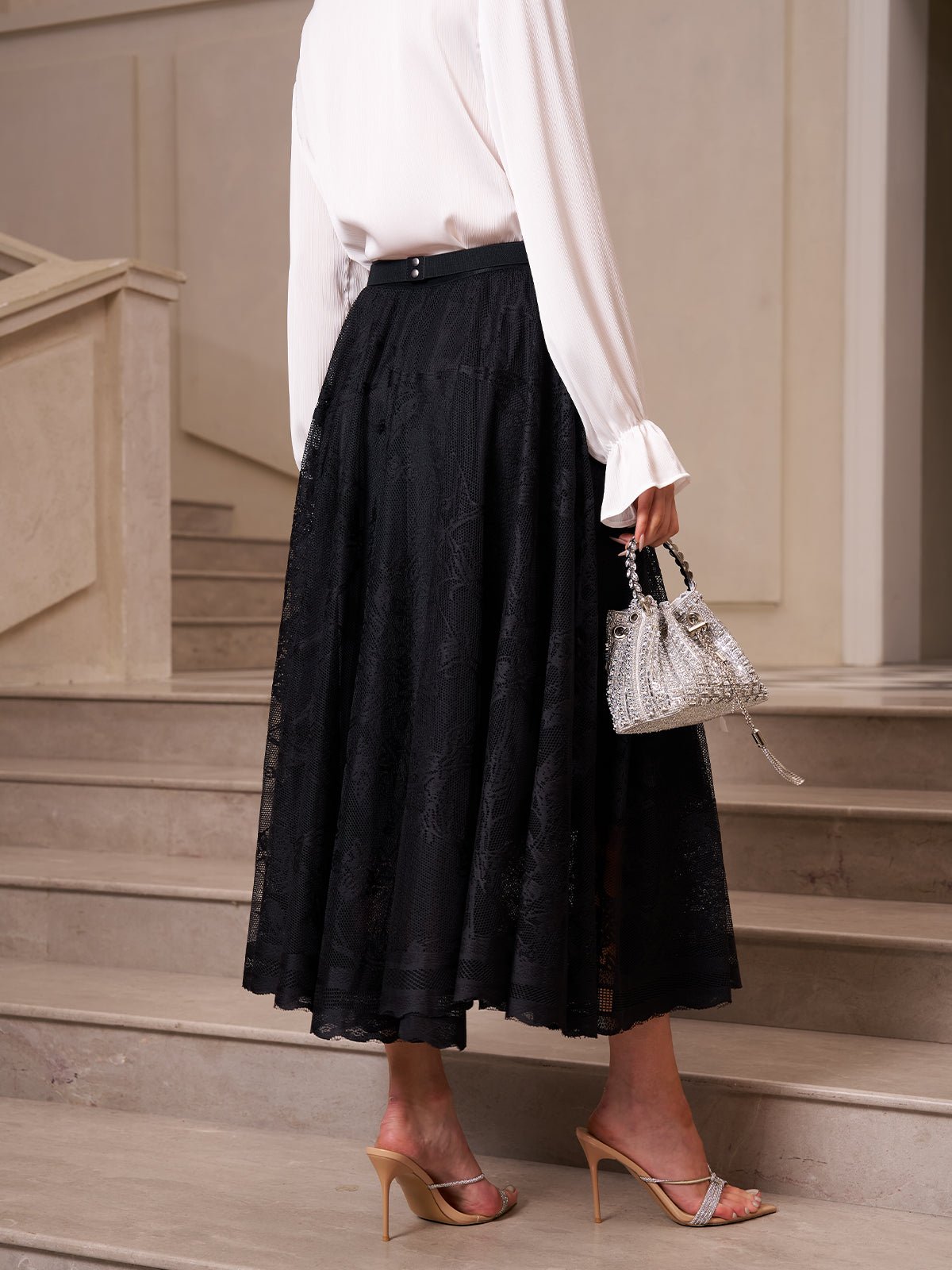 A person stands on a staircase holding a small silver handbag, dressed in a white blouse, beige heeled sandals, and the Social Queen Jacquard Lace A-Line Midi Skirt in black.