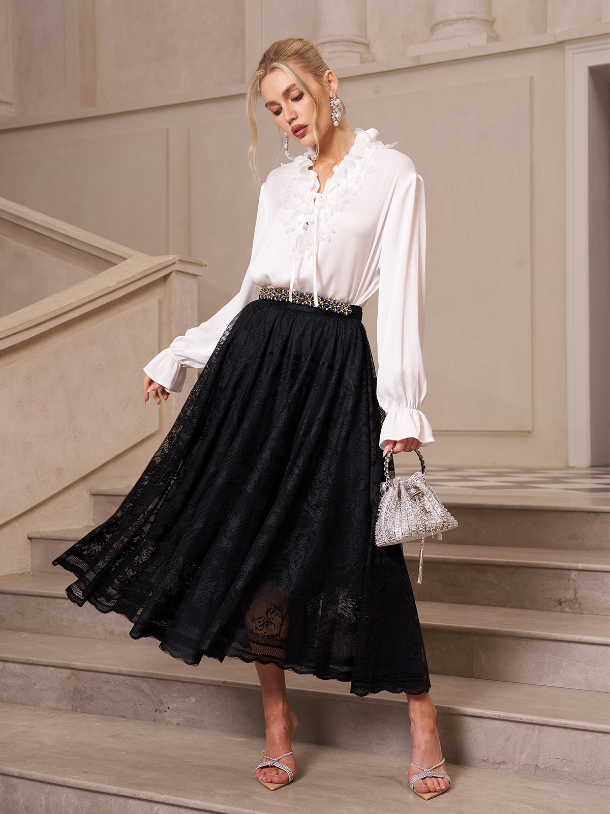 A woman stands on stairs in a long-sleeved white blouse, silver heels, and a Jacquard Lace A-Line Midi Skirt by Social Queen, holding a small silver handbag. Light-colored walls and stairs form the background.